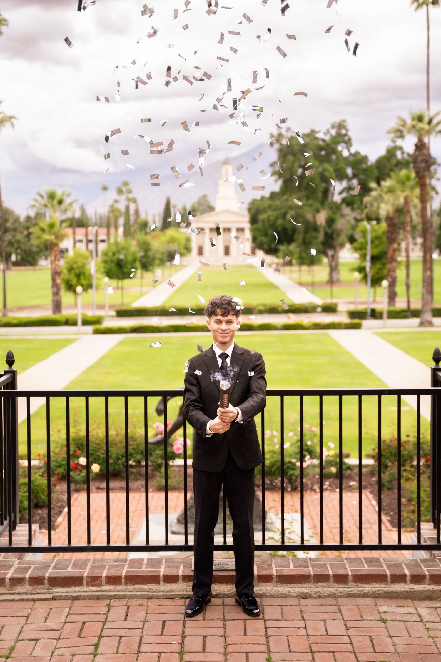 Young man in black suit pops confetti cannon at scenic university garden graduation photo.