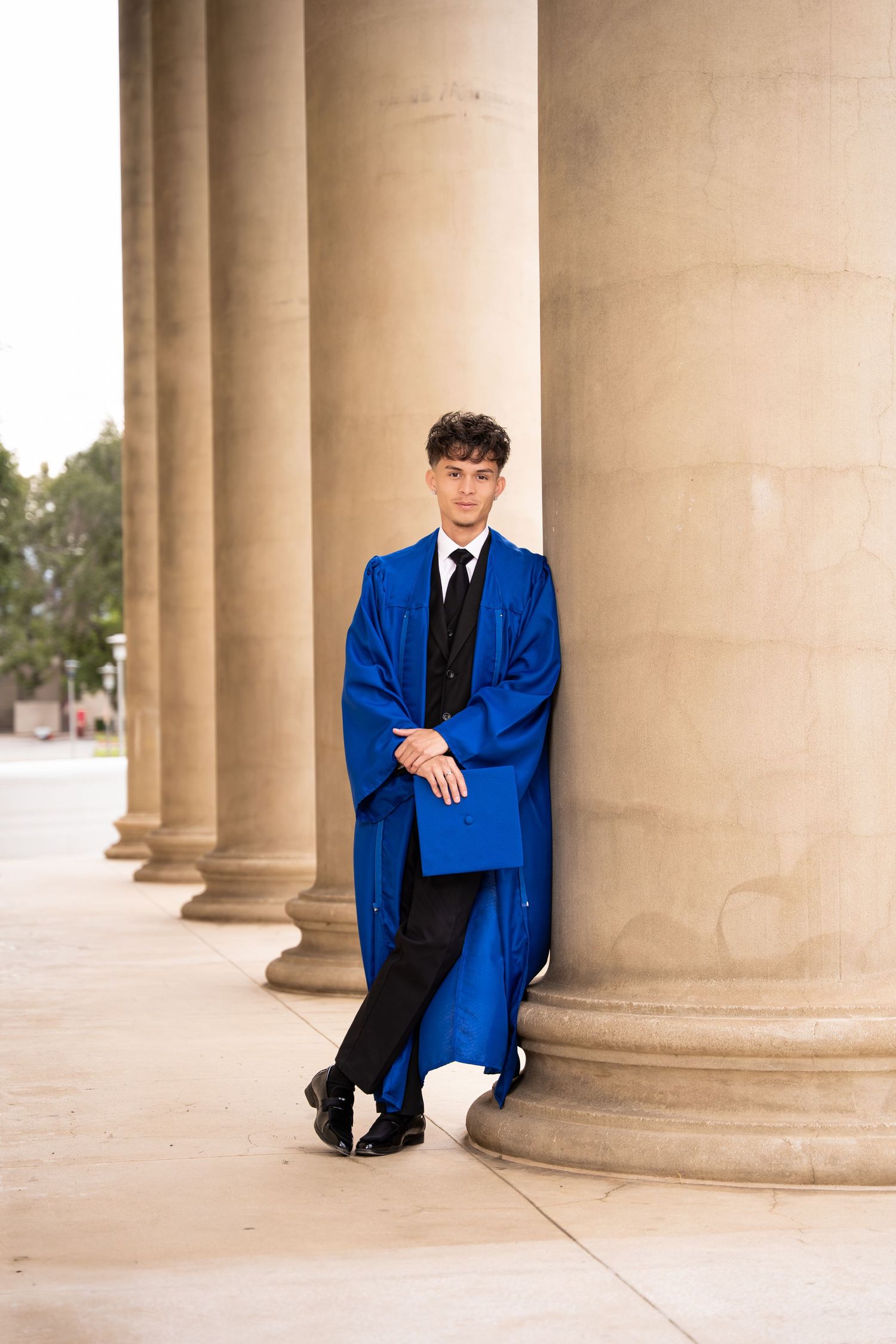 Male graduate in royal blue graduation gown leaning against stone columns in outdoor corridor.
