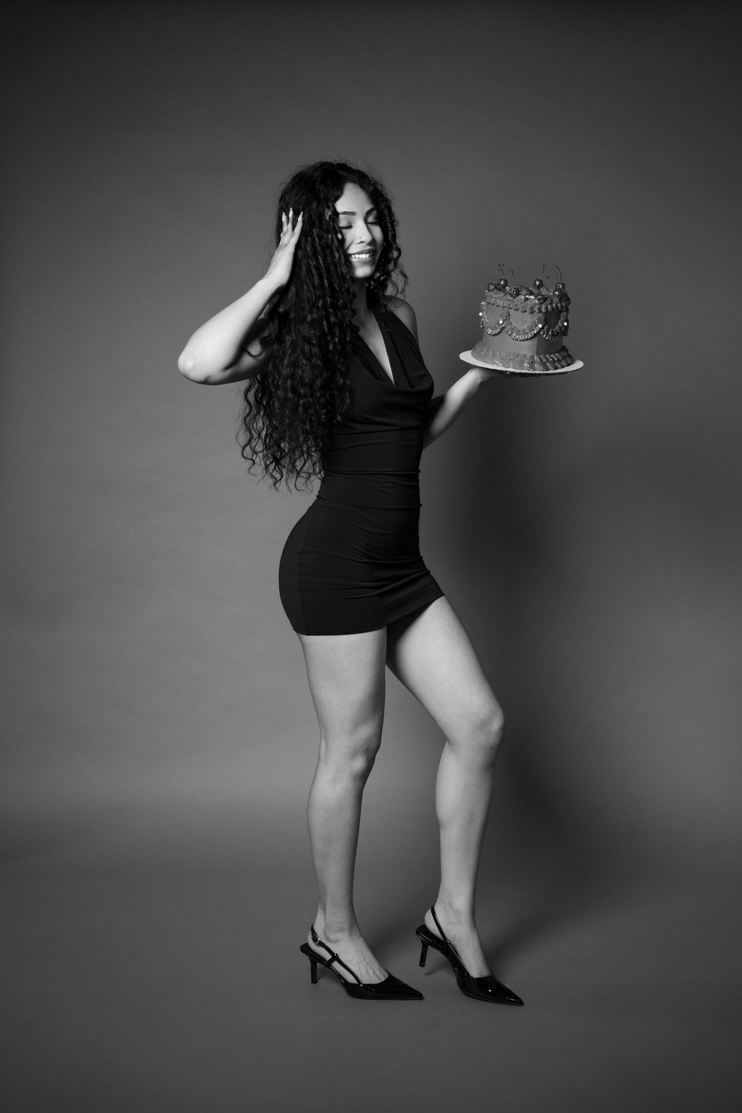 Woman in black mini dress and heels holding birthday cake, striking a pose in a black and white studio photo.