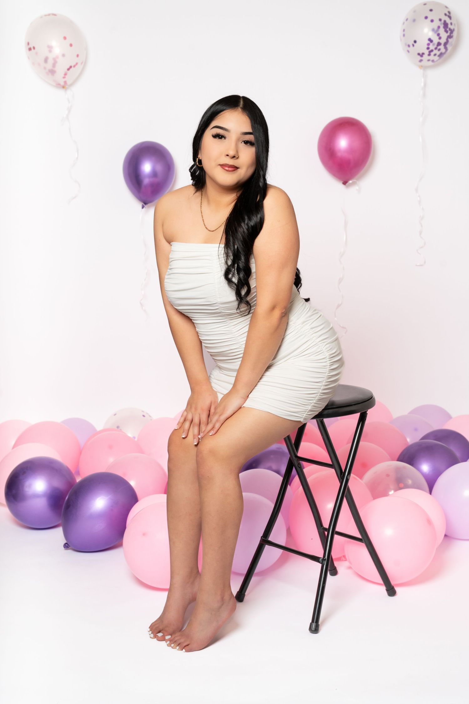 Young woman in white ruched dress sitting on stool surrounded by pink and purple balloons on white background.