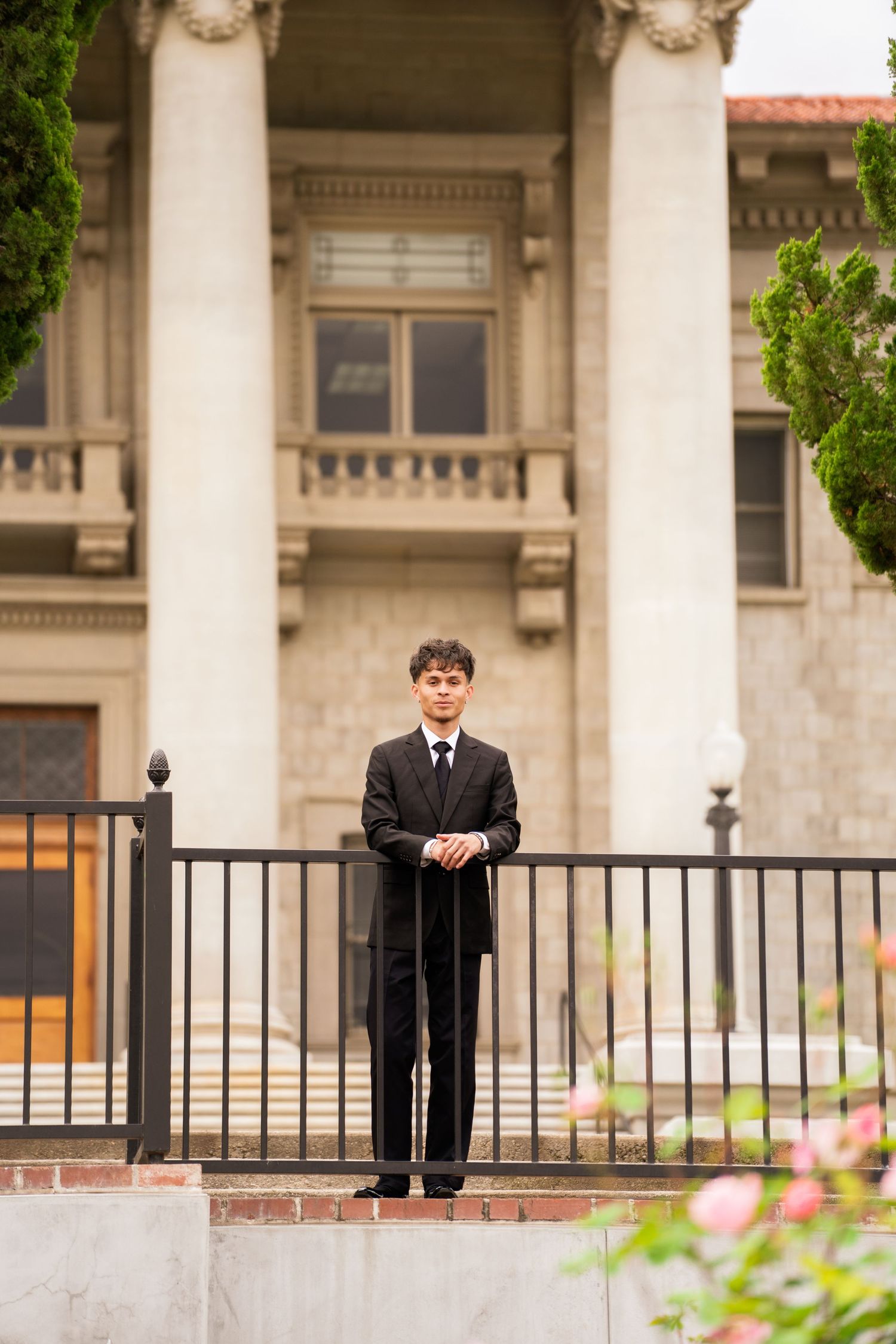 Young man in black suit standing at iron fence in front of grand columned building exterior.