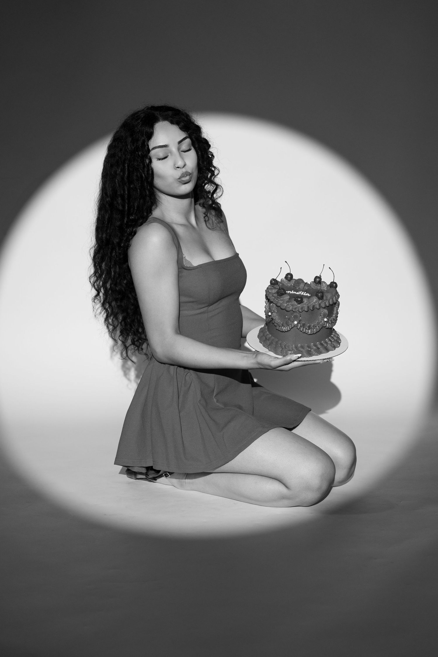Black and white photo of woman in dress kneeling holding decorated cake in spotlight.