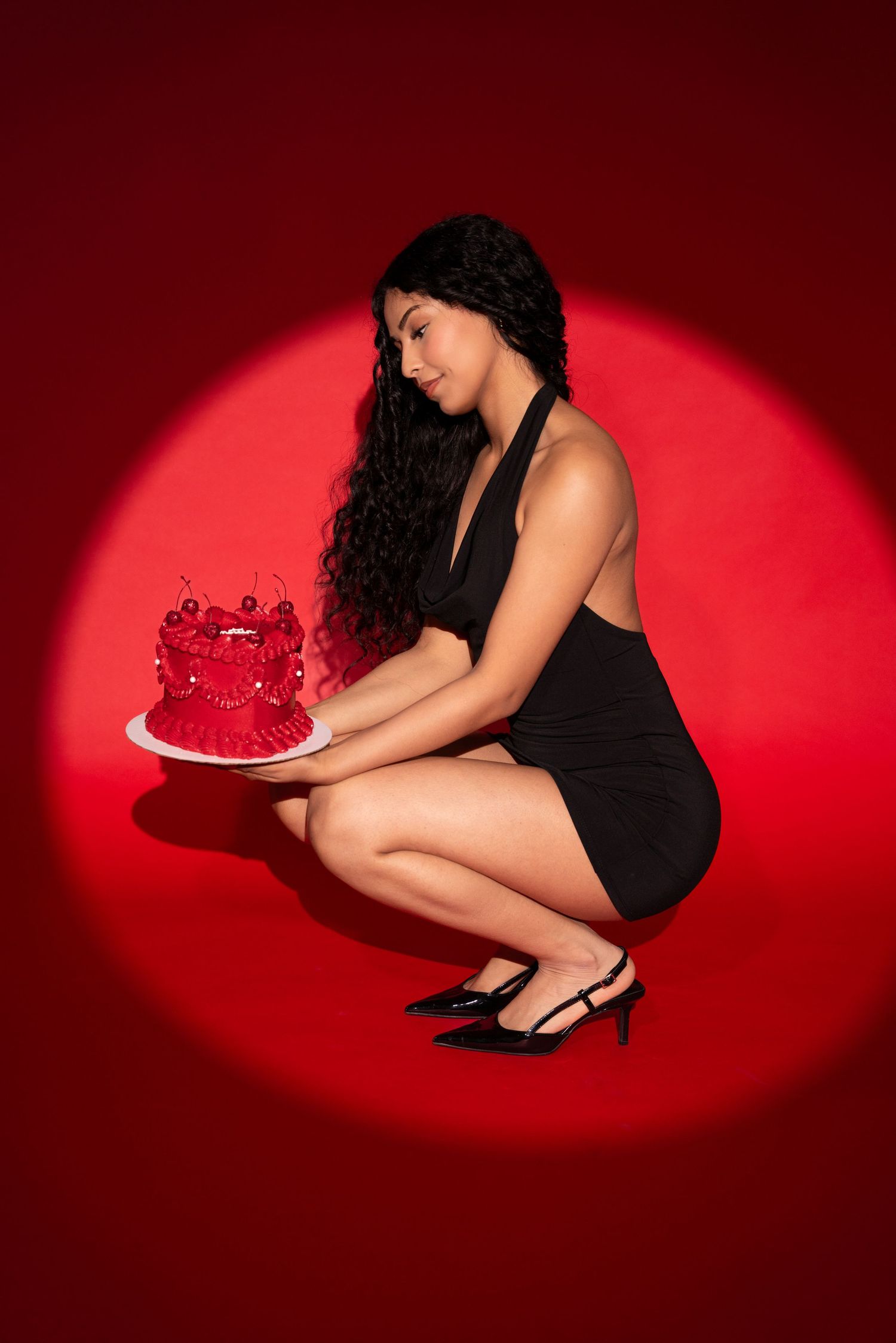 Woman in black dress crouching holding red decorated cake against red spotlight backdrop.
