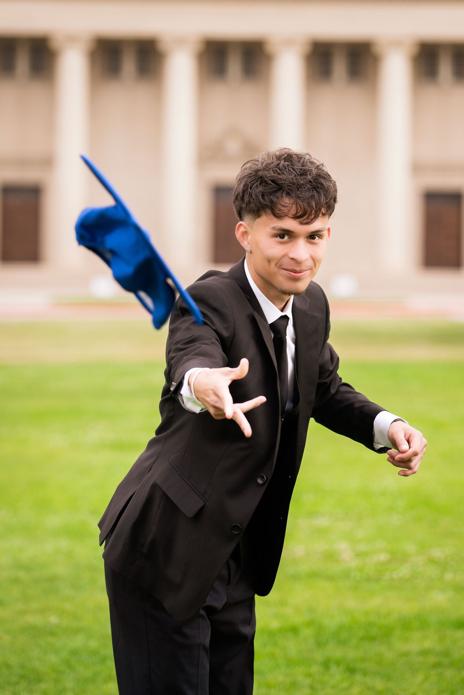 Young man in black suit tossing blue graduation cap on green lawn near columned building.