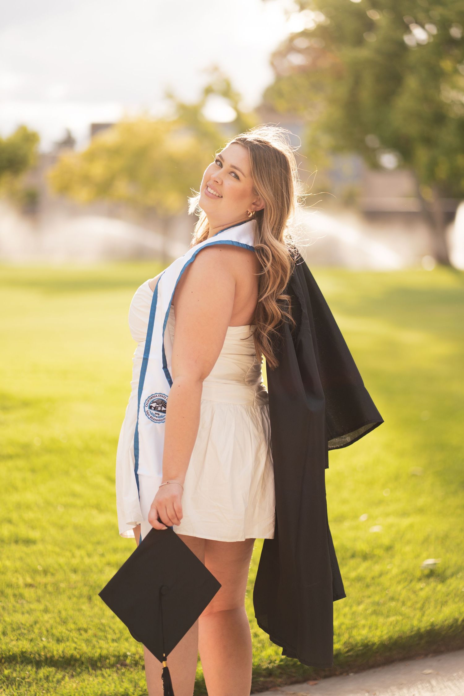 Smiling female graduate in white dress holds black cap and gown in sunny park setting.