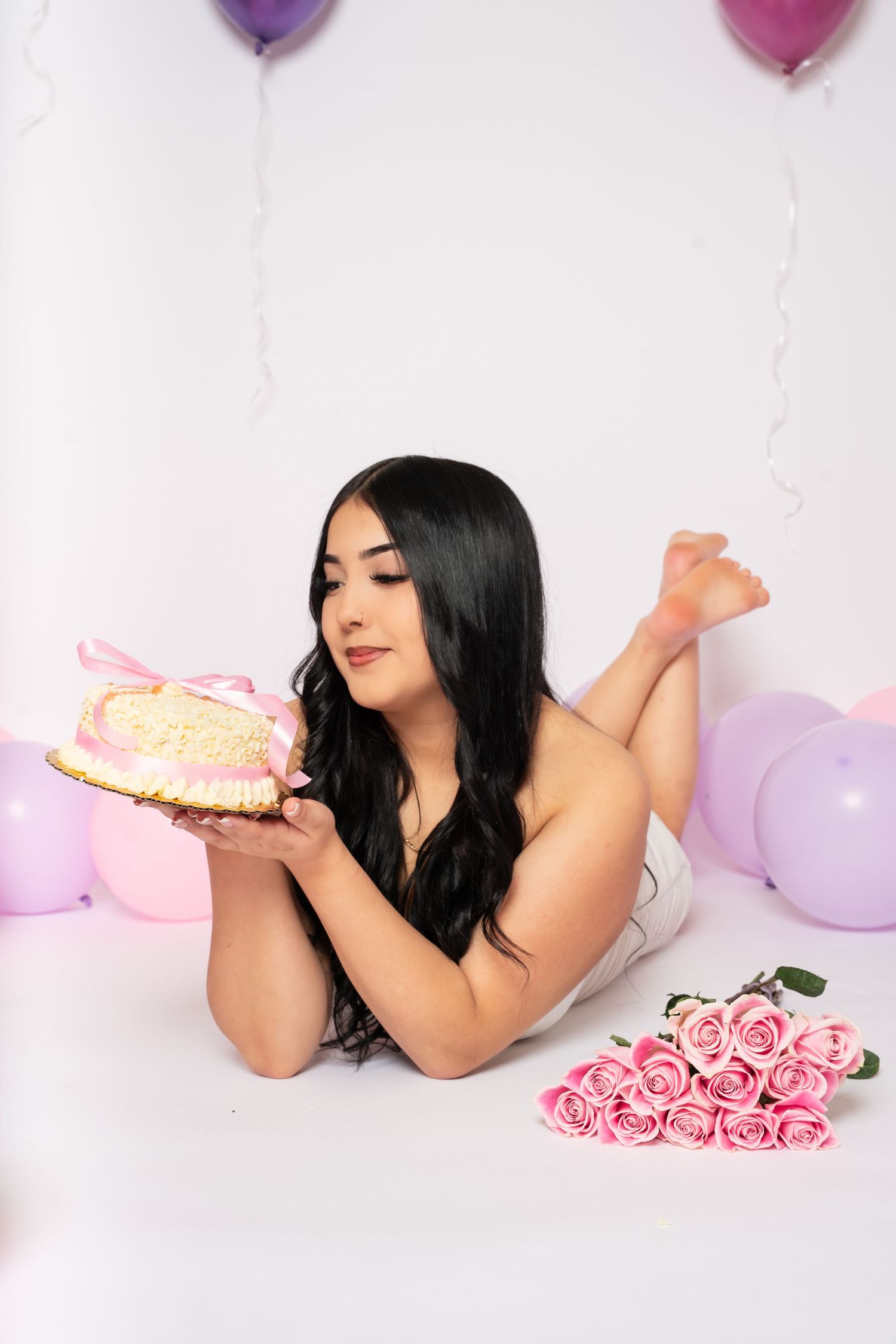 Young woman lying on floor holding birthday cake with pink roses and colorful balloons in background.