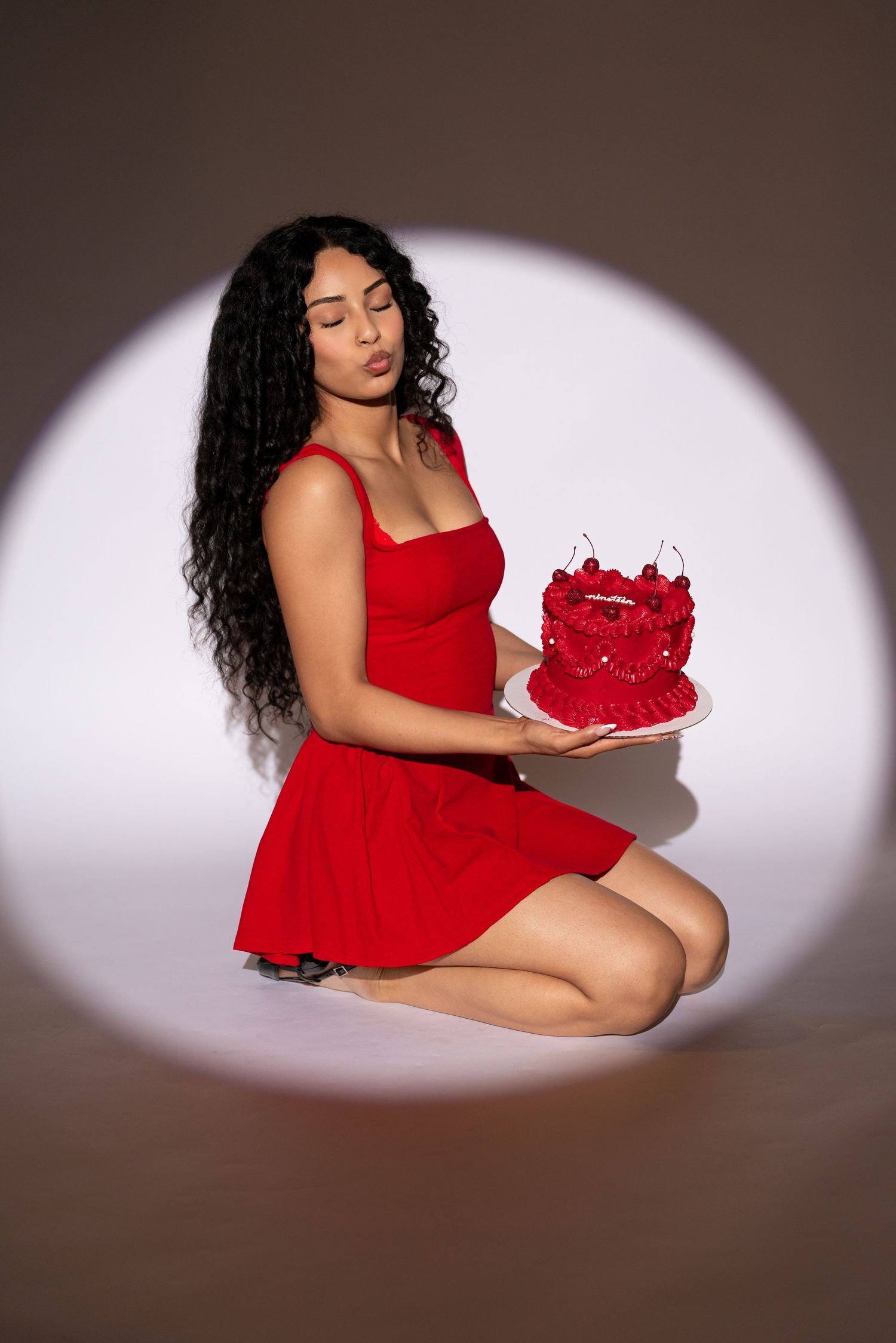 Woman in red dress kneeling holding red decorated cake in spotlight circle.