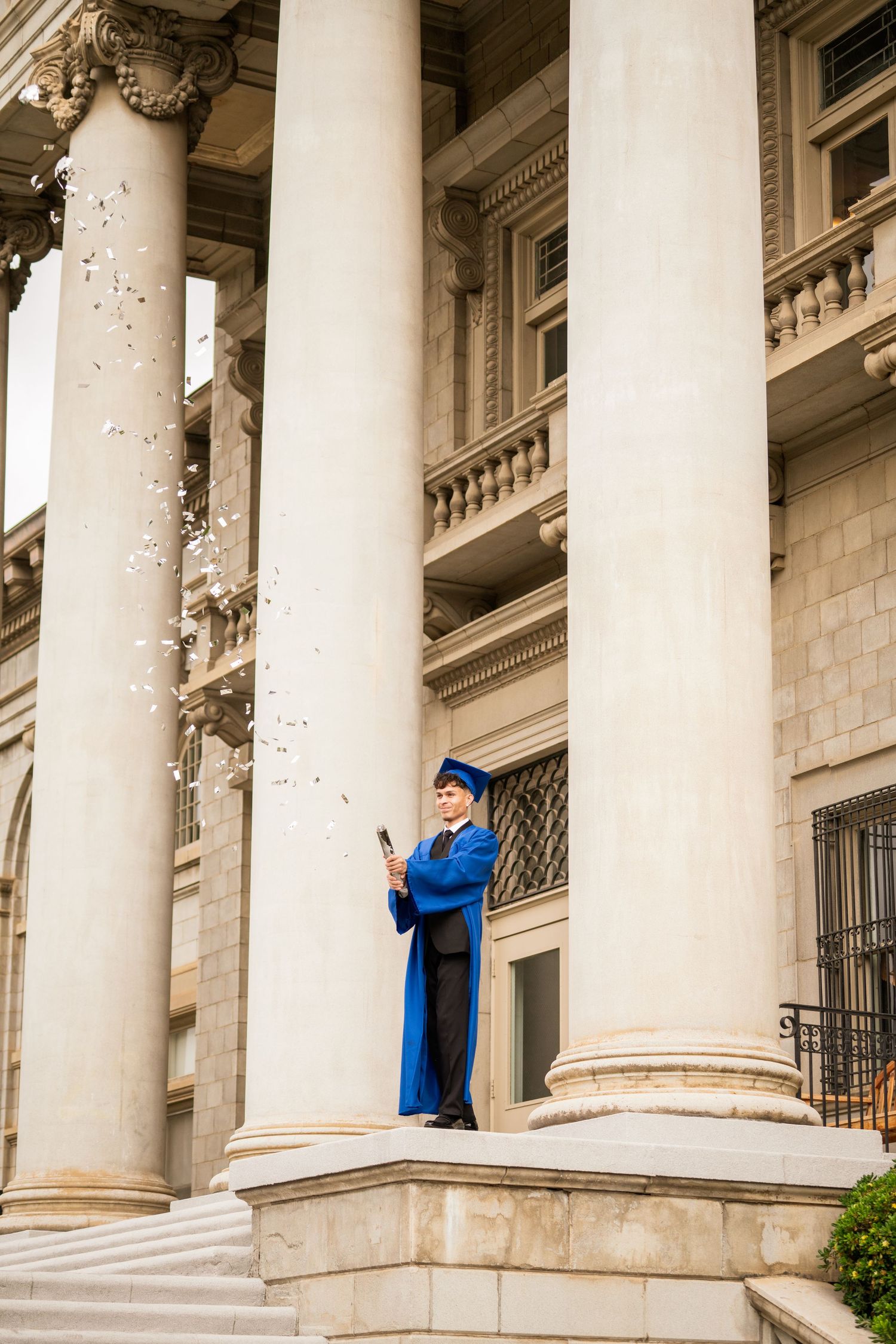 Graduate in blue cap and gown shooting confetti popper between large columns of historic building.