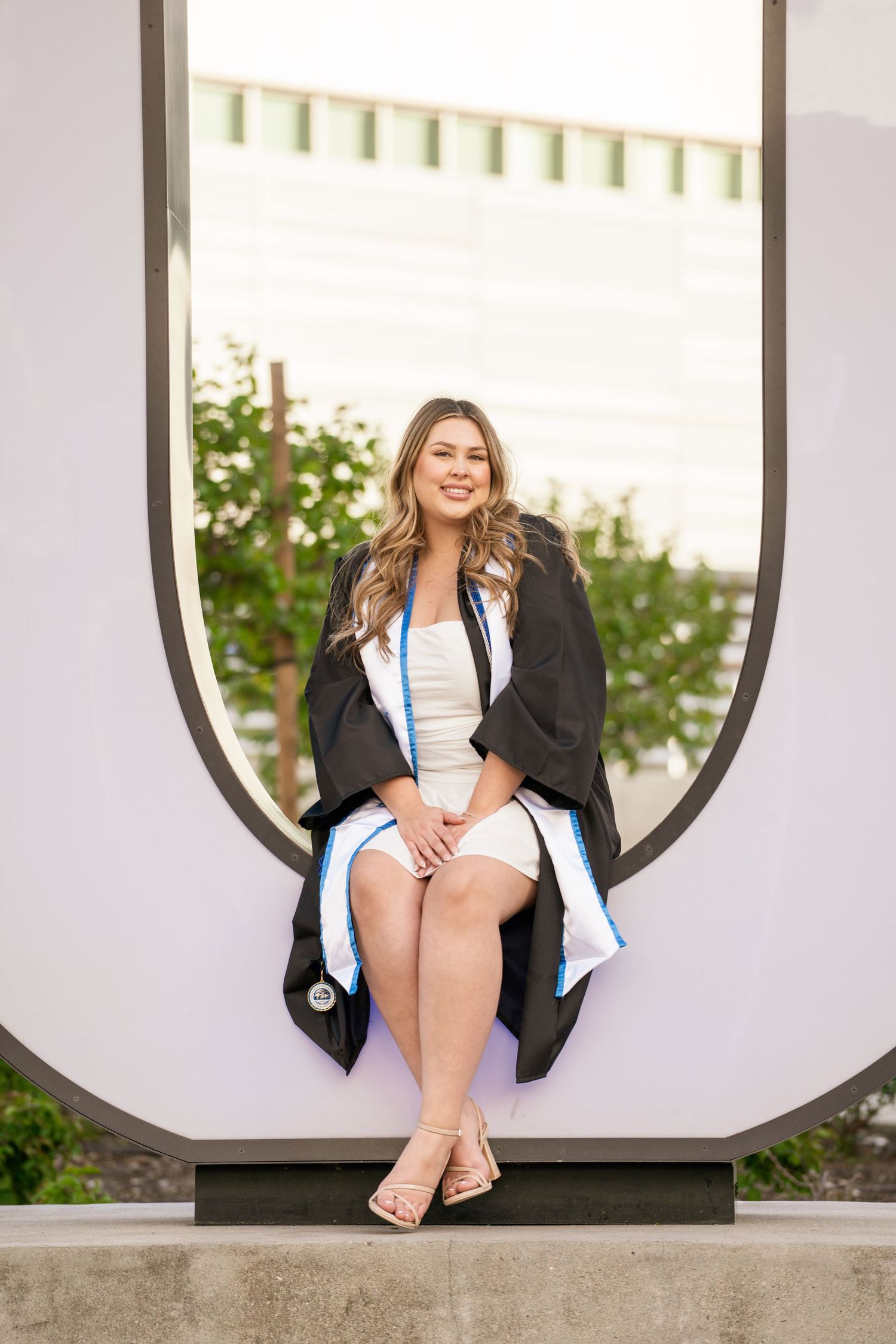 Female graduate in black gown sits inside circular campus sculpture for graduation portrait.