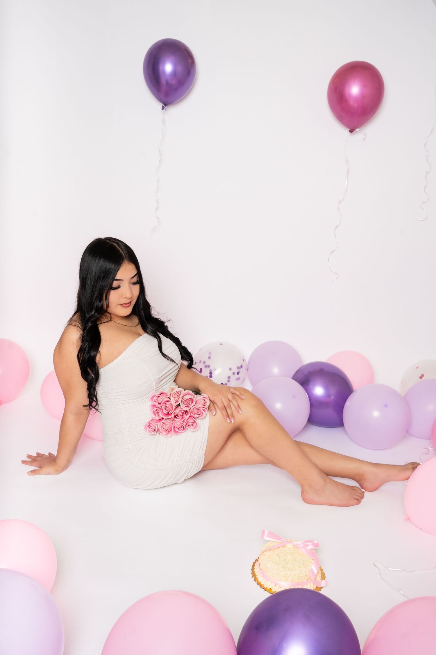 Young woman in white dress sitting on floor with pink roses and colorful balloons in background.