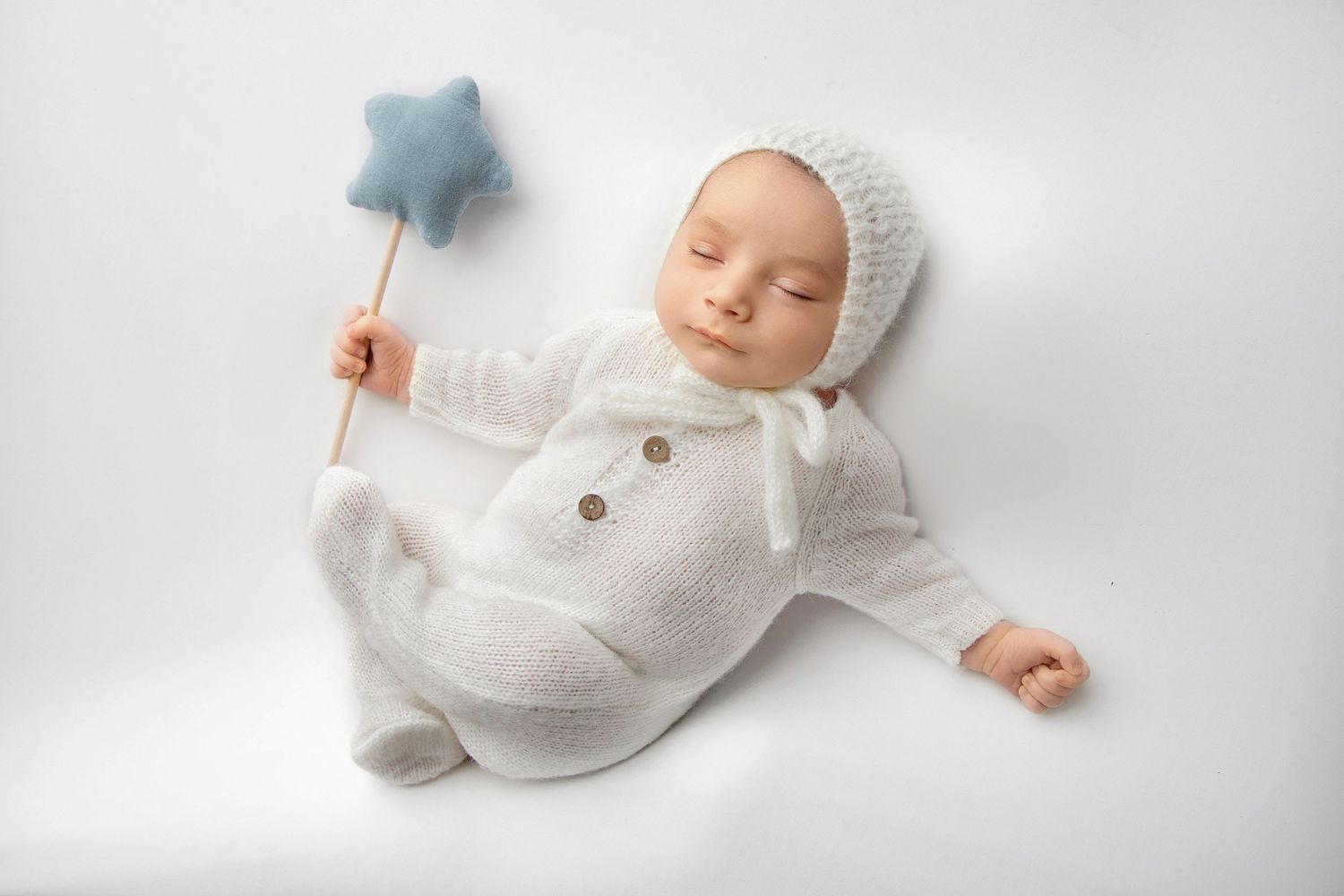 Sleeping newborn baby in a soft white knit outfit and bonnet, posed on a neutral background during a Lakewood NJ newborn photography session