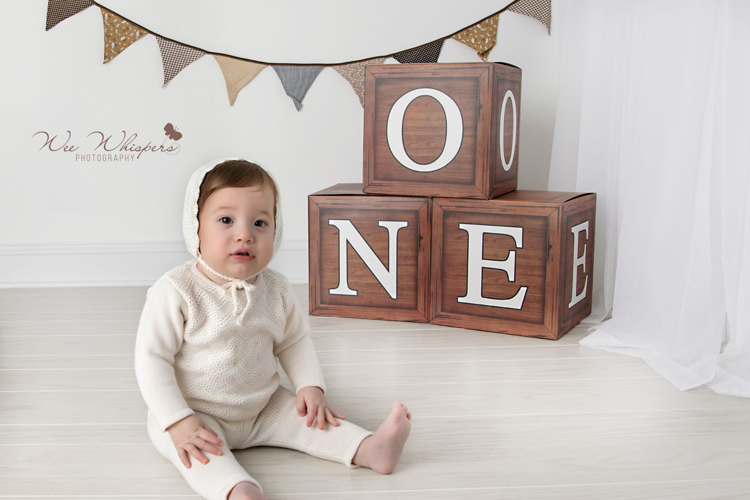 A baby sitting on the floor, wearing a white knitted outfit and bonnet. The child is surrounded by large wooden blocks spelling 