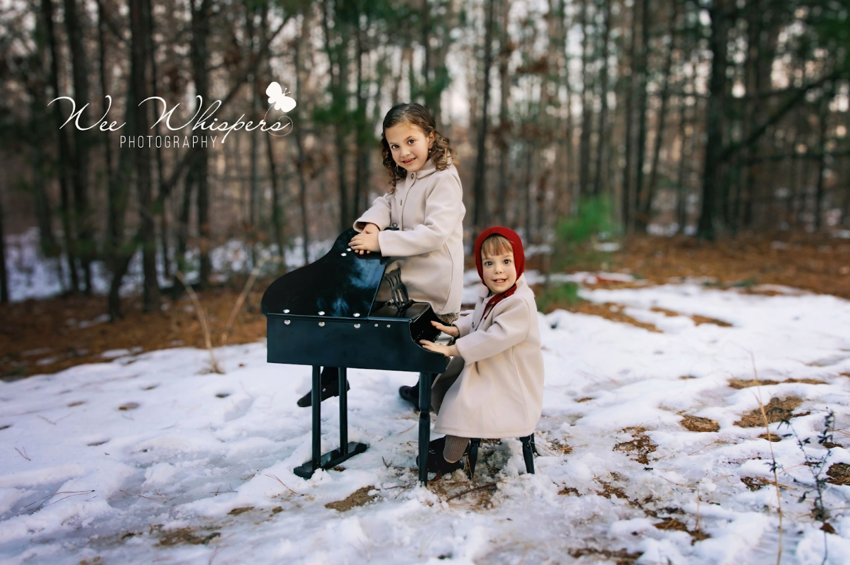 Two young children in light coats pose outdoors in the snow beside a small black toy piano in a wooded area.