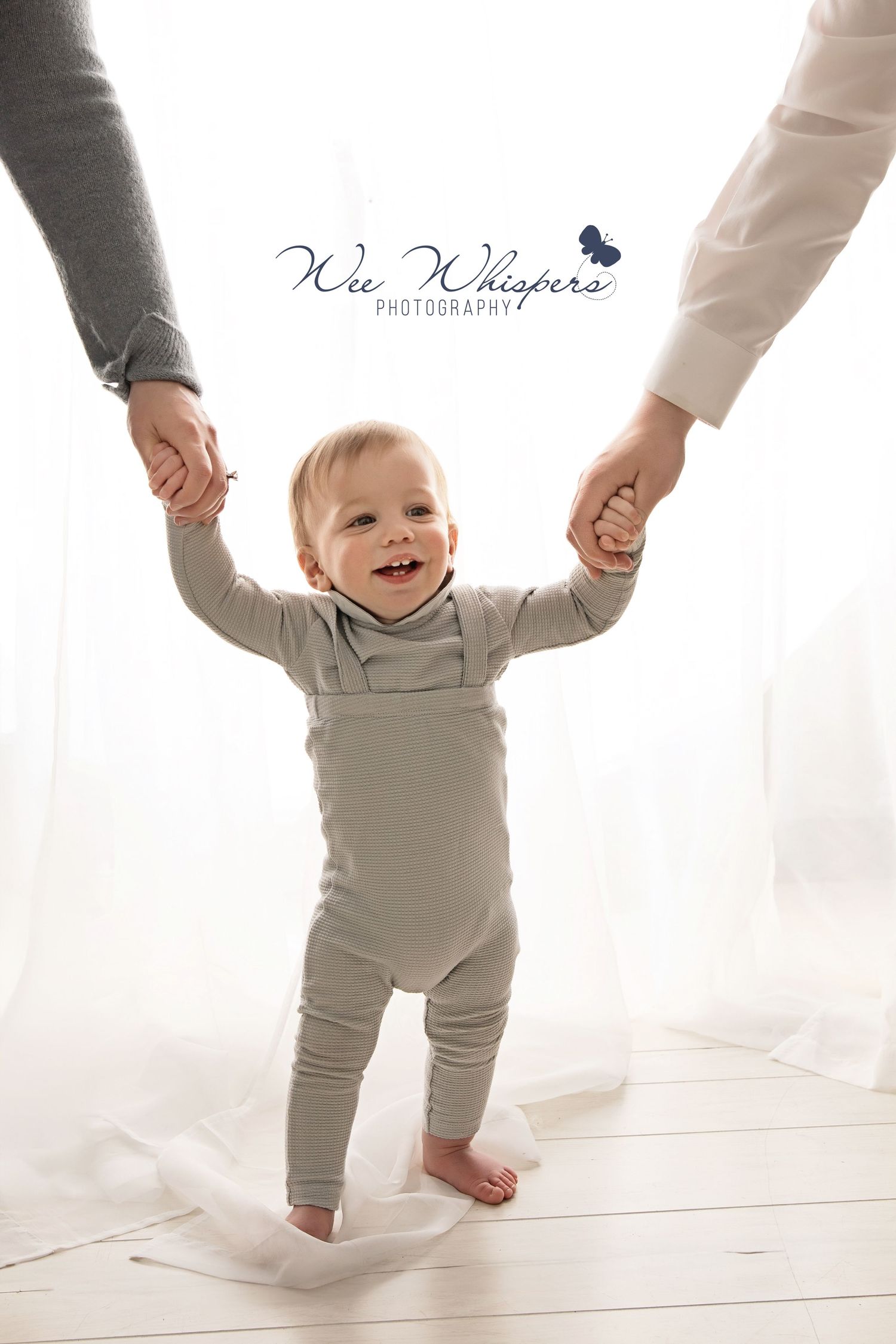 Baby in grey outfit learning to walk while holding hands in bright white studio.