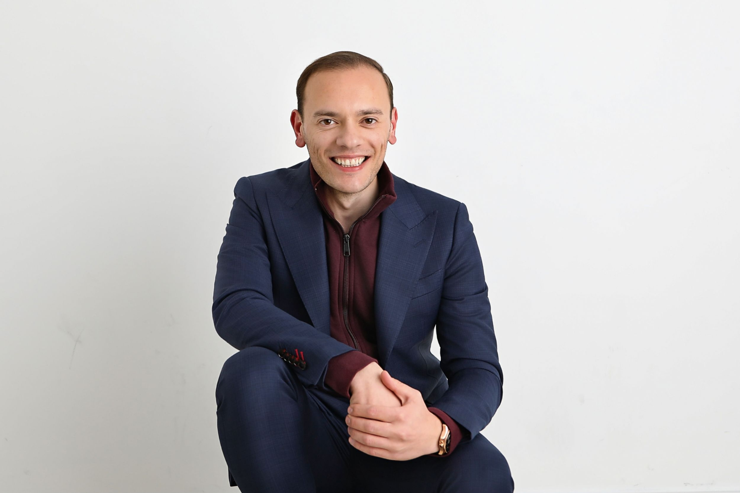 Man in a dark suit and maroon sweater smiling, seated against a plain white background.