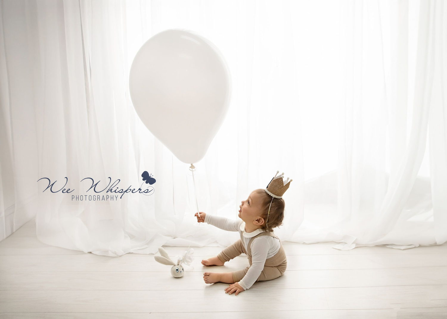 Toddler wearing a crown and holding a white balloon in a bright studio