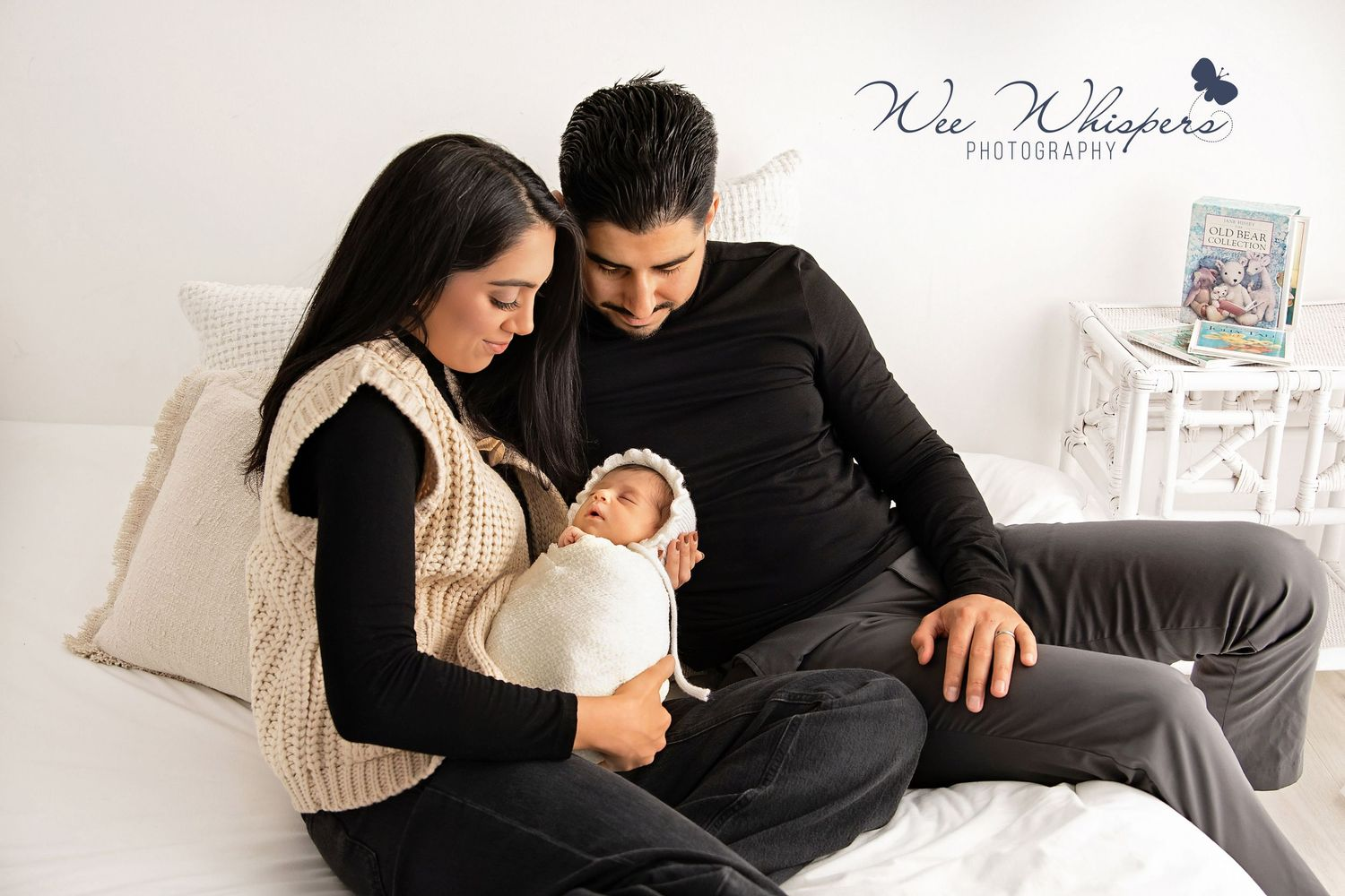 Parents sitting on a bed, looking lovingly at their swaddled newborn.