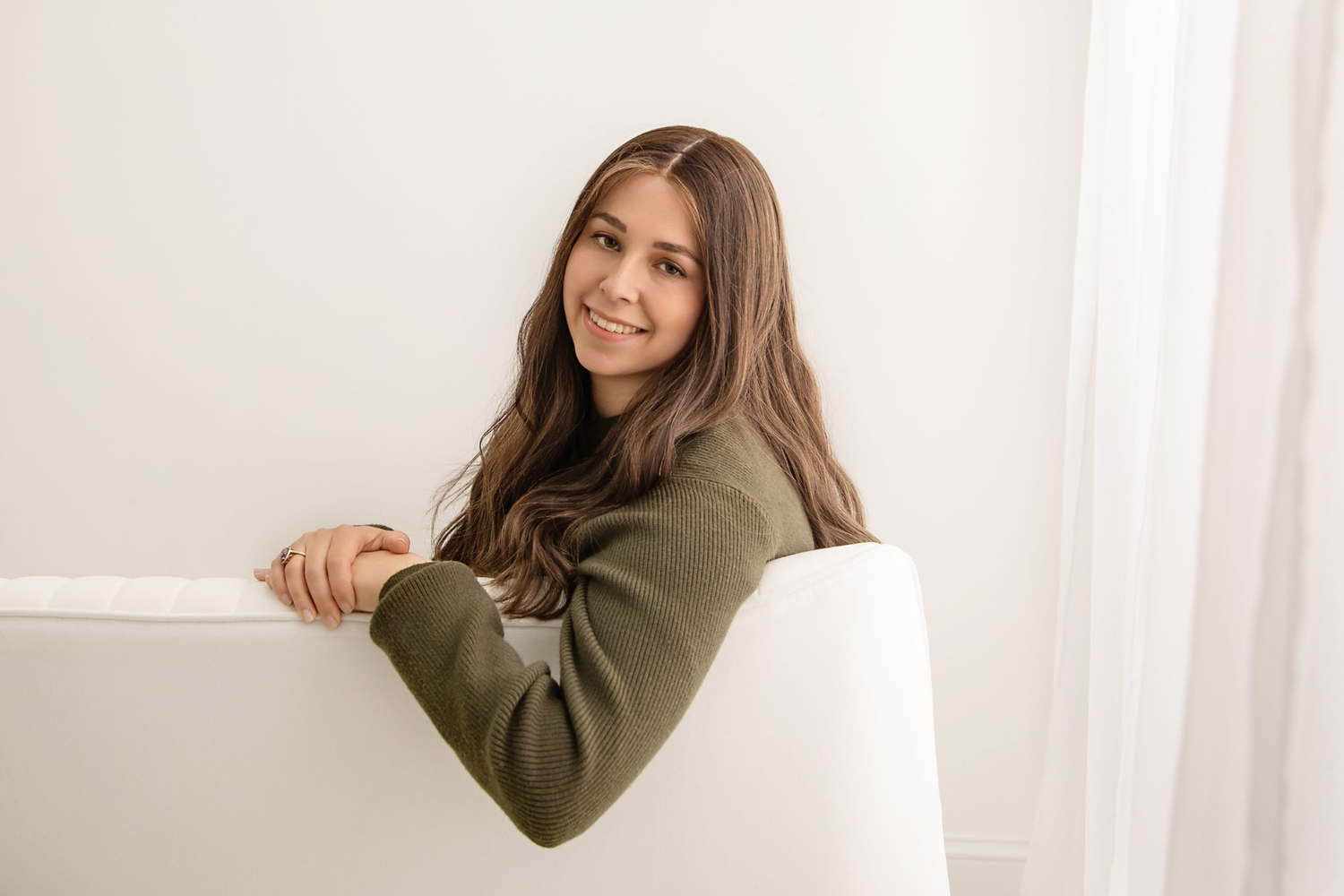 Woman in an olive green sweater leaning on a white couch, smiling at the camera.