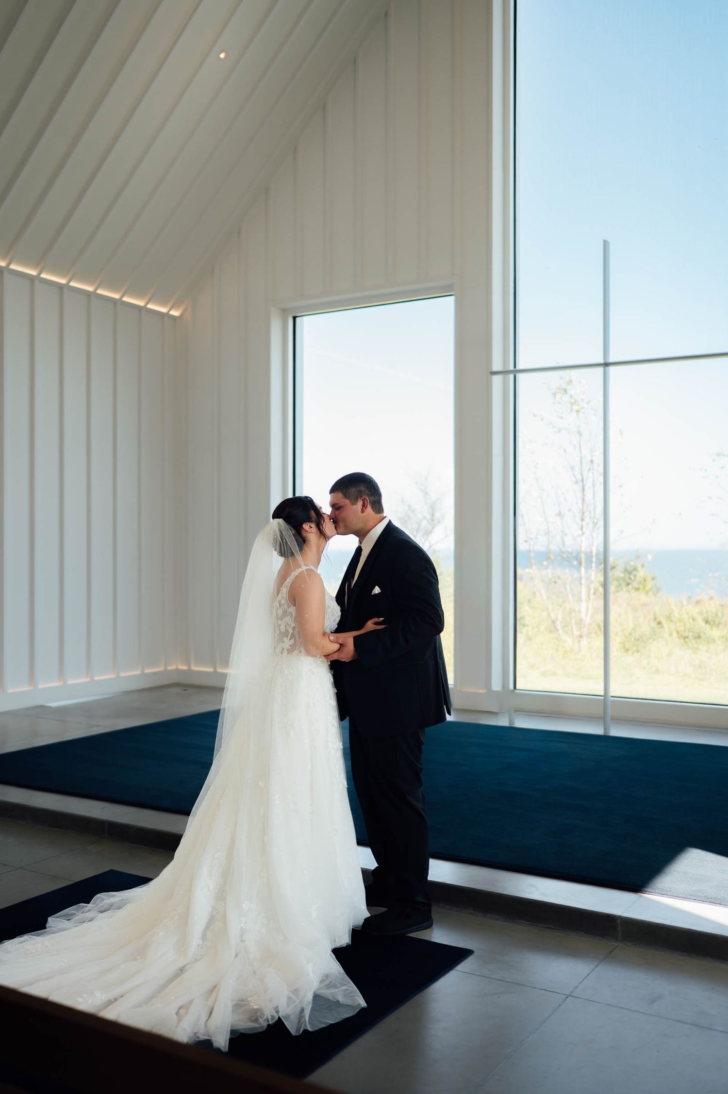 Bride and groom share a first kiss in a modern white chapel with floor-to-ceiling windows overlooking the ocean.