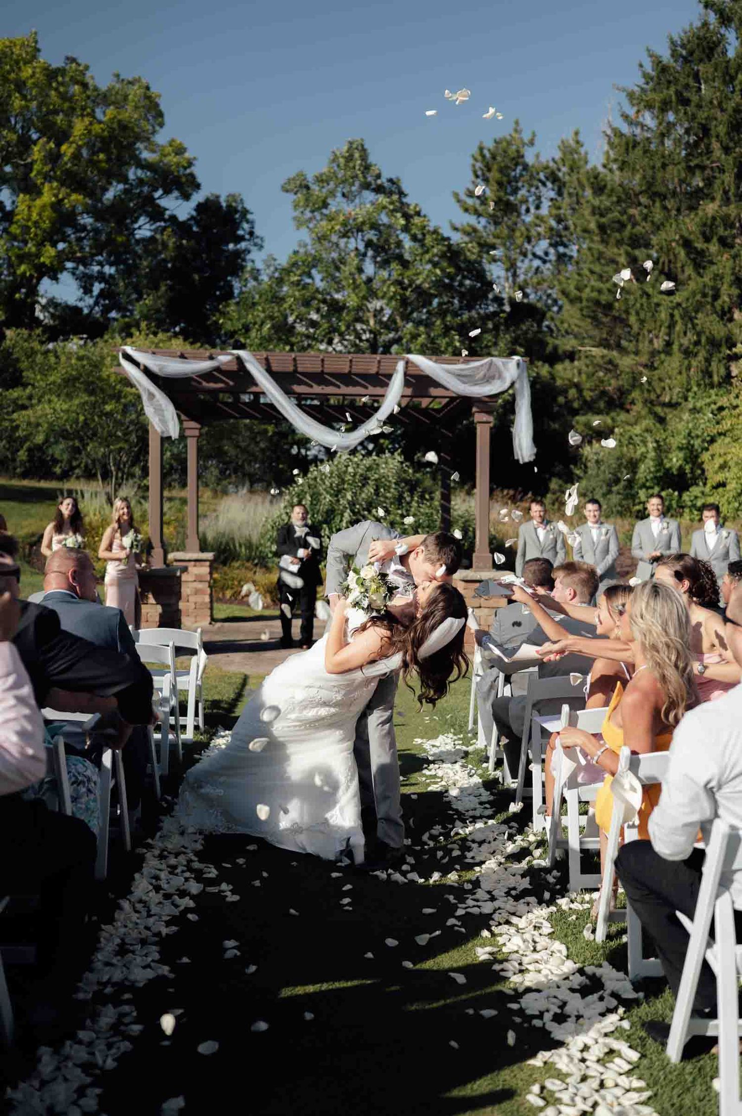 Bride and groom share a kiss at outdoor wedding ceremony as guests toss white rose petals along the aisle.