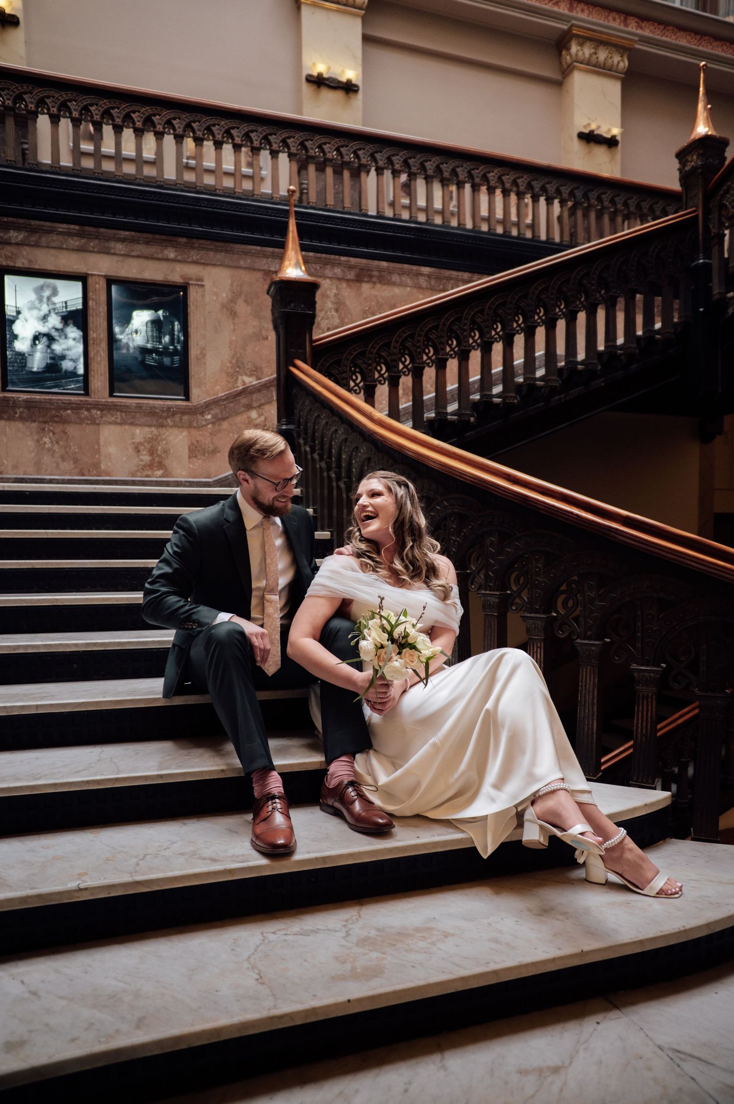 Bride and groom laughing together on ornate wooden staircase, bride holding bouquet in elegant white dress.