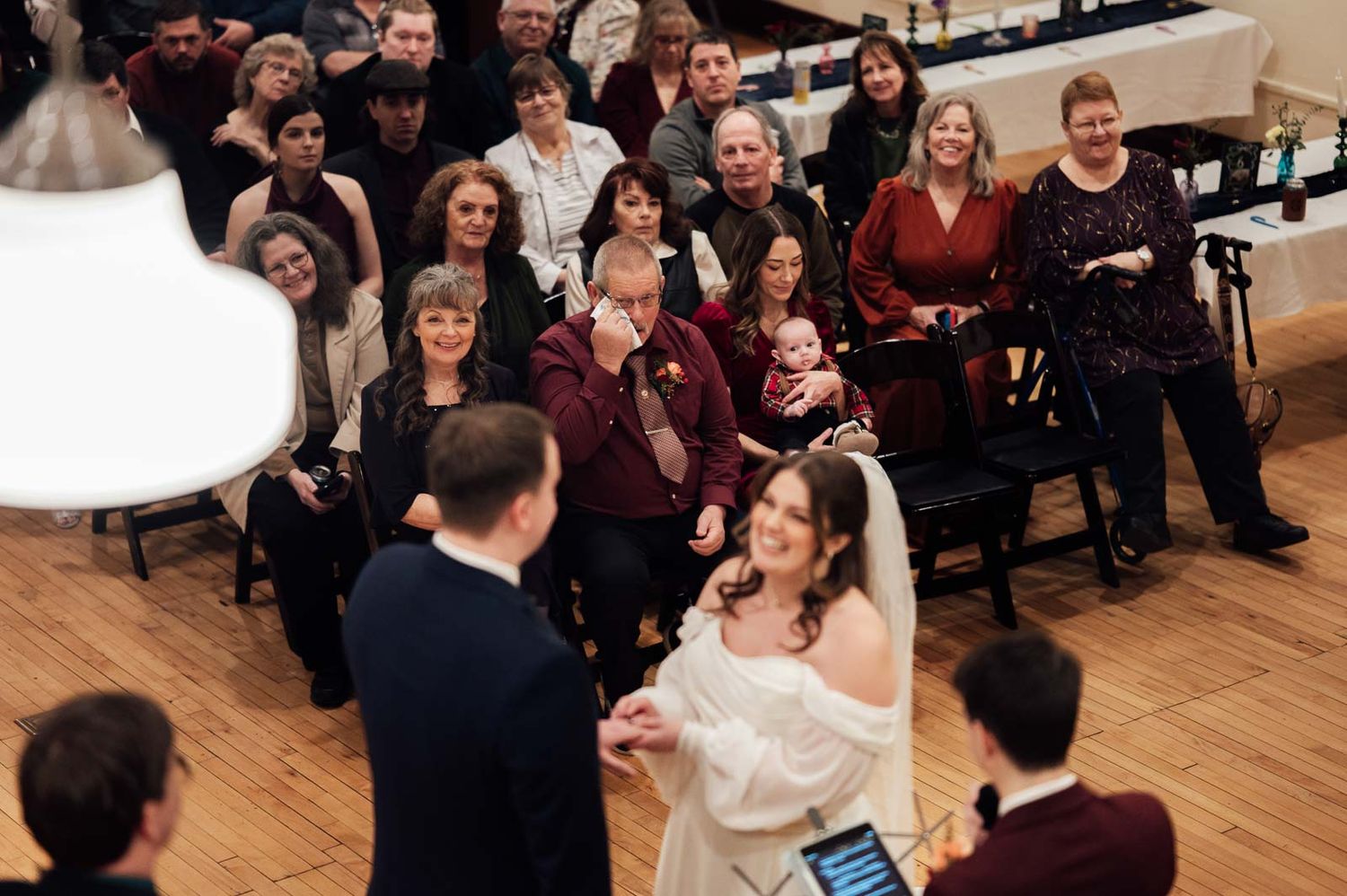 Bride and groom exchange vows at indoor wedding ceremony with guests seated in rows behind them.