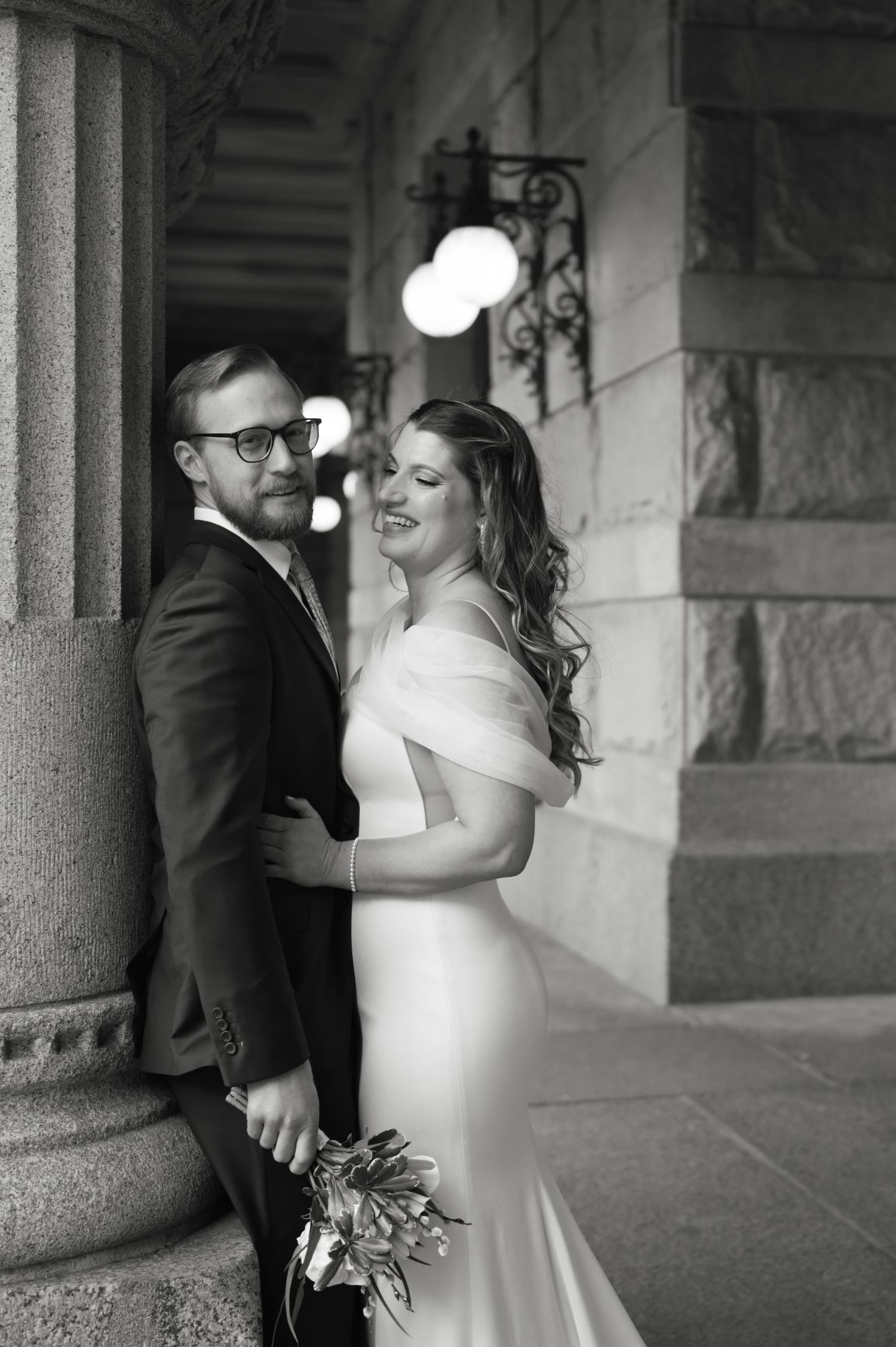 Black and white photo of a smiling bride in off-shoulder gown and groom in suit posing by stone columns.