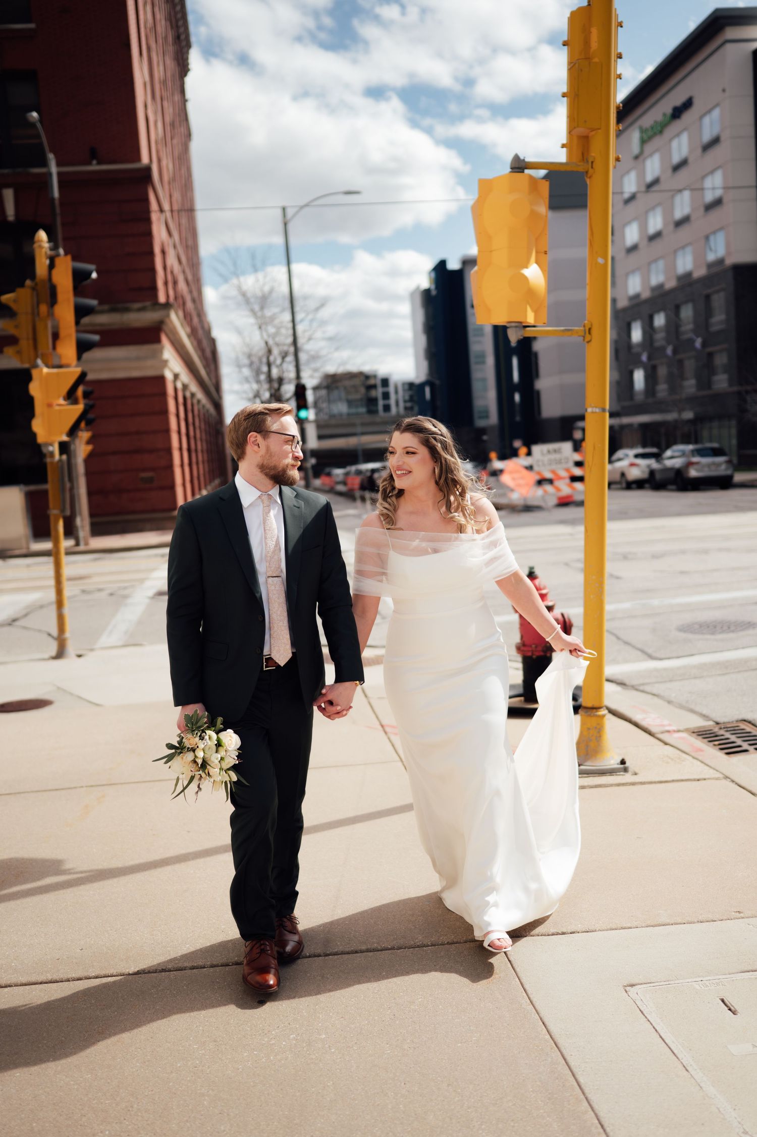 Bride and groom walk hand-in-hand on a sunny urban sidewalk, she in a white gown, he in a black suit with bouquet.