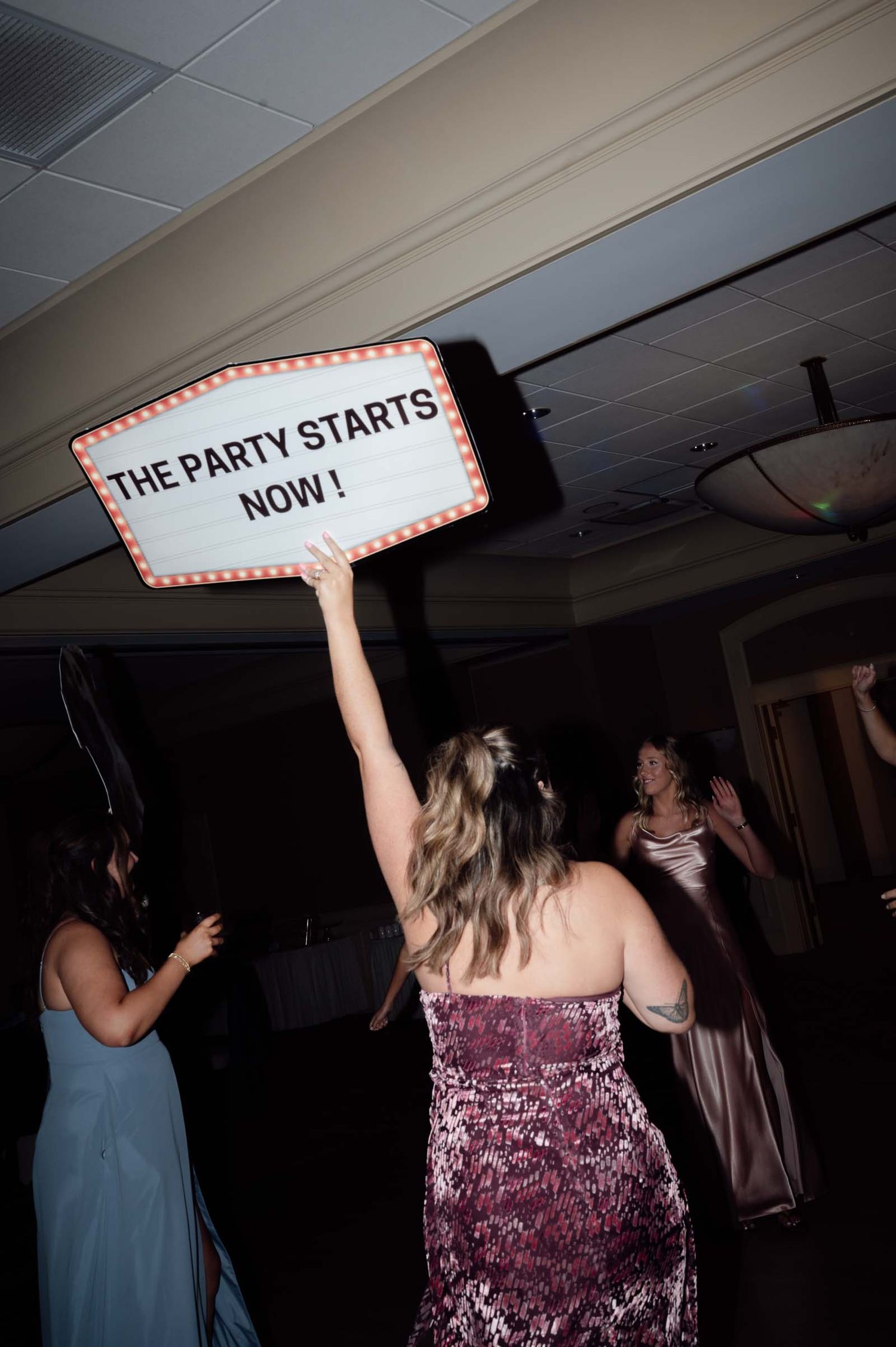 Women dancing at a wedding reception, one holding a lit marquee sign reading The Party Starts Now.