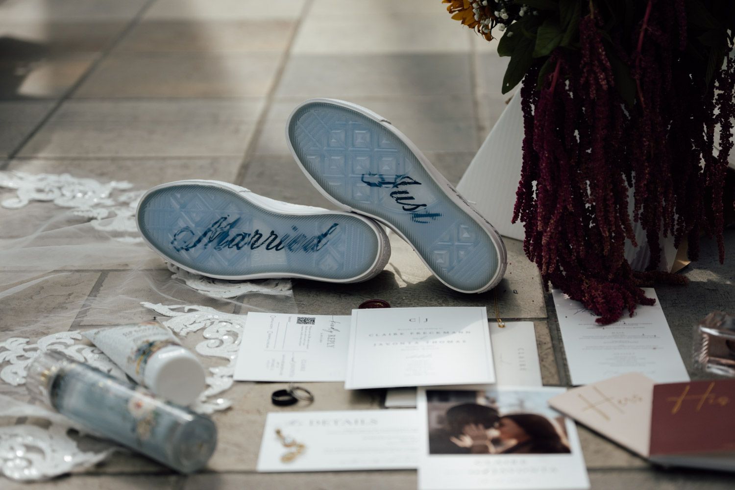Wedding flat lay featuring Just Married sneakers, invitations, rings, and burgundy floral bouquet on tile floor.