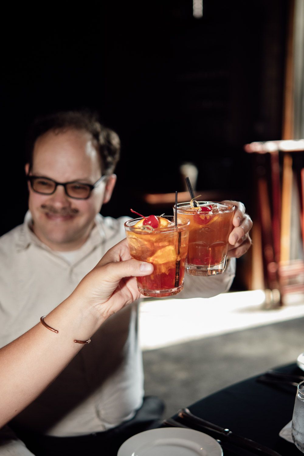 Man in glasses smiling as two hands clink orange cocktails with cherries at a restaurant table.