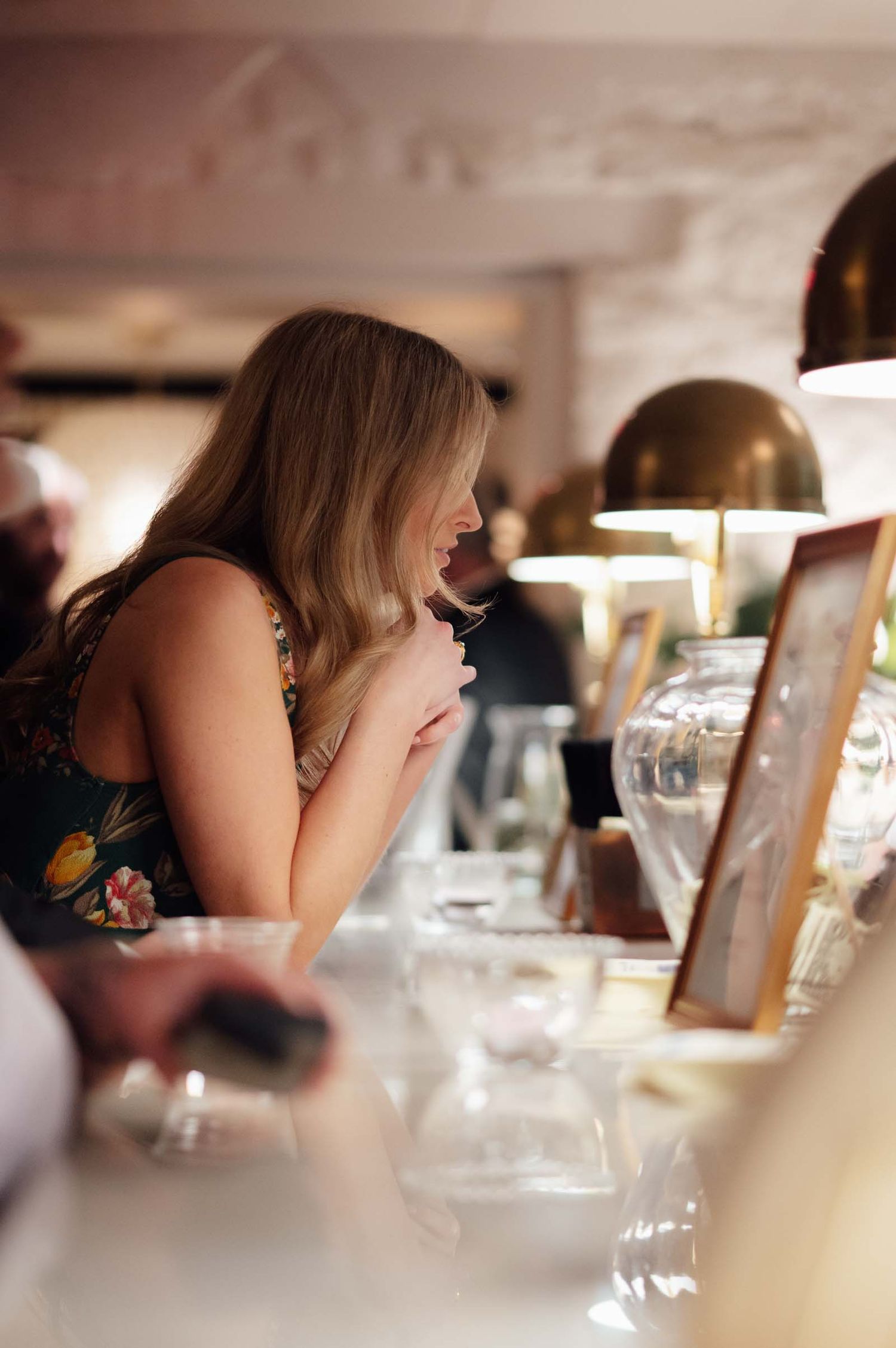 A woman with long blonde hair smiles while browsing items on a display table at an elegant indoor market or boutique.