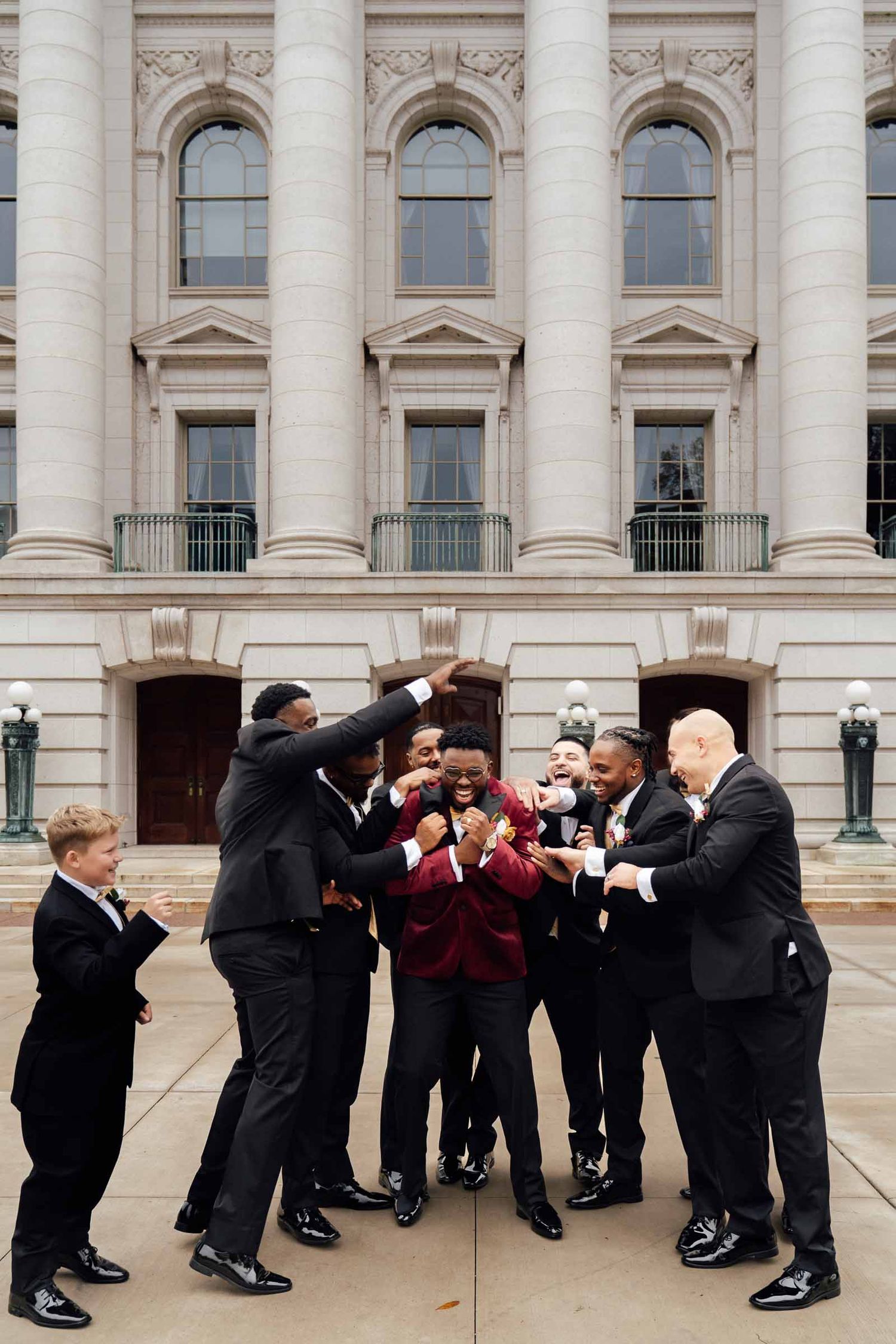 Groomsmen in black tuxedos celebrate with groom in burgundy jacket outside grand neoclassical building.