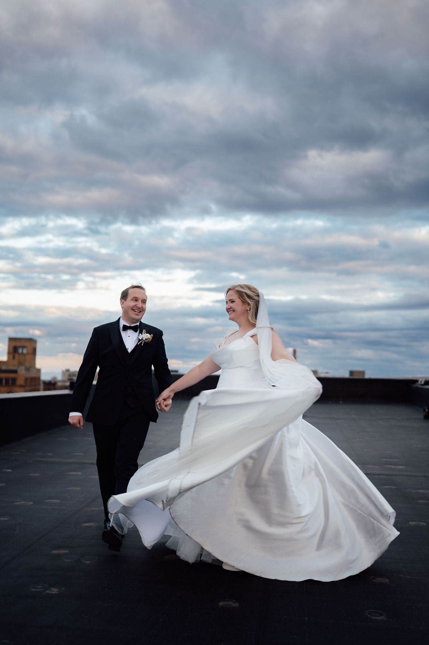 Bride in flowing white gown twirls with groom in black tuxedo on rooftop under dramatic cloudy sky.
