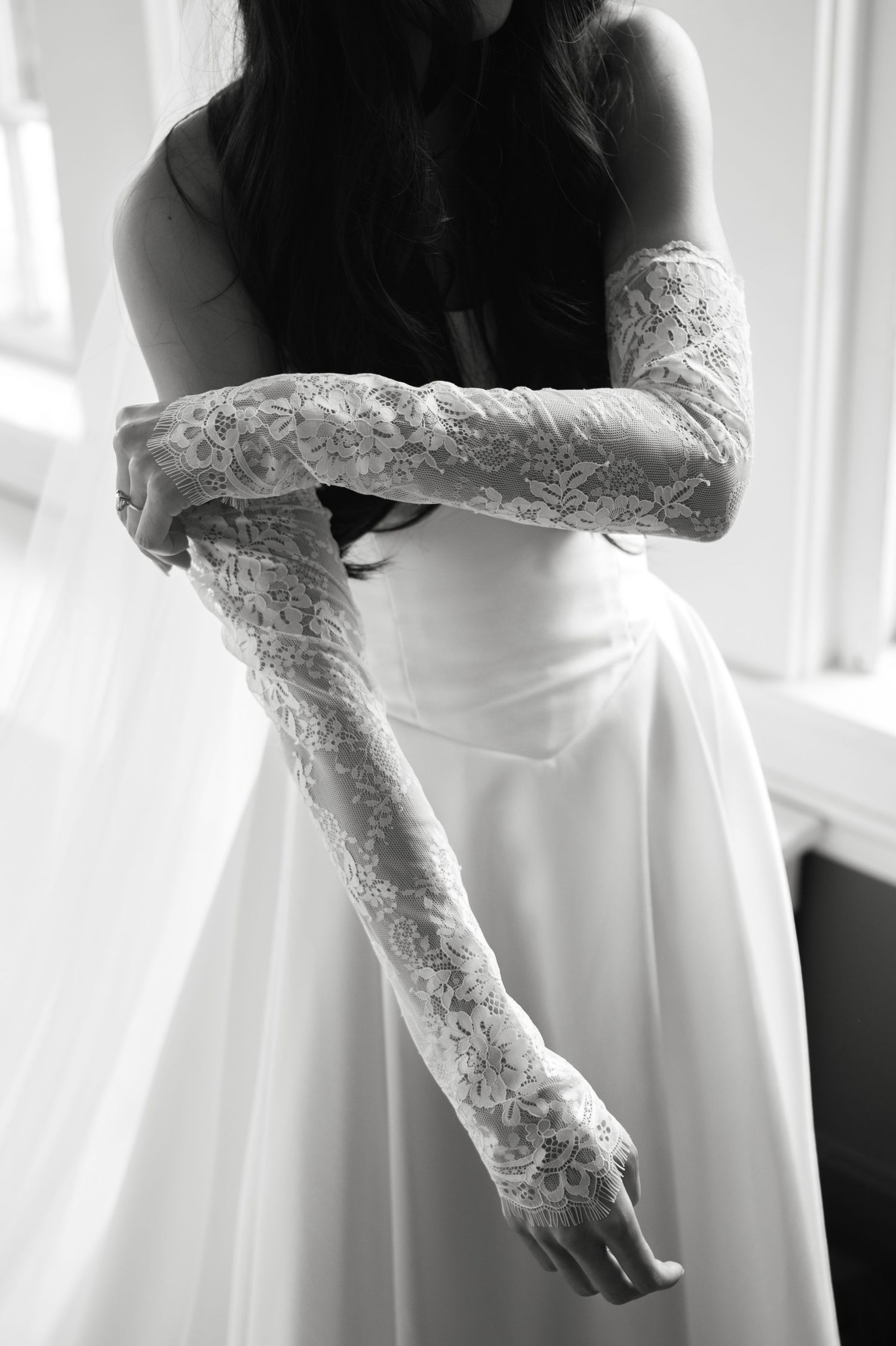 Bride in white satin gown adjusting elegant lace elbow-length gloves, black and white wedding photography.
