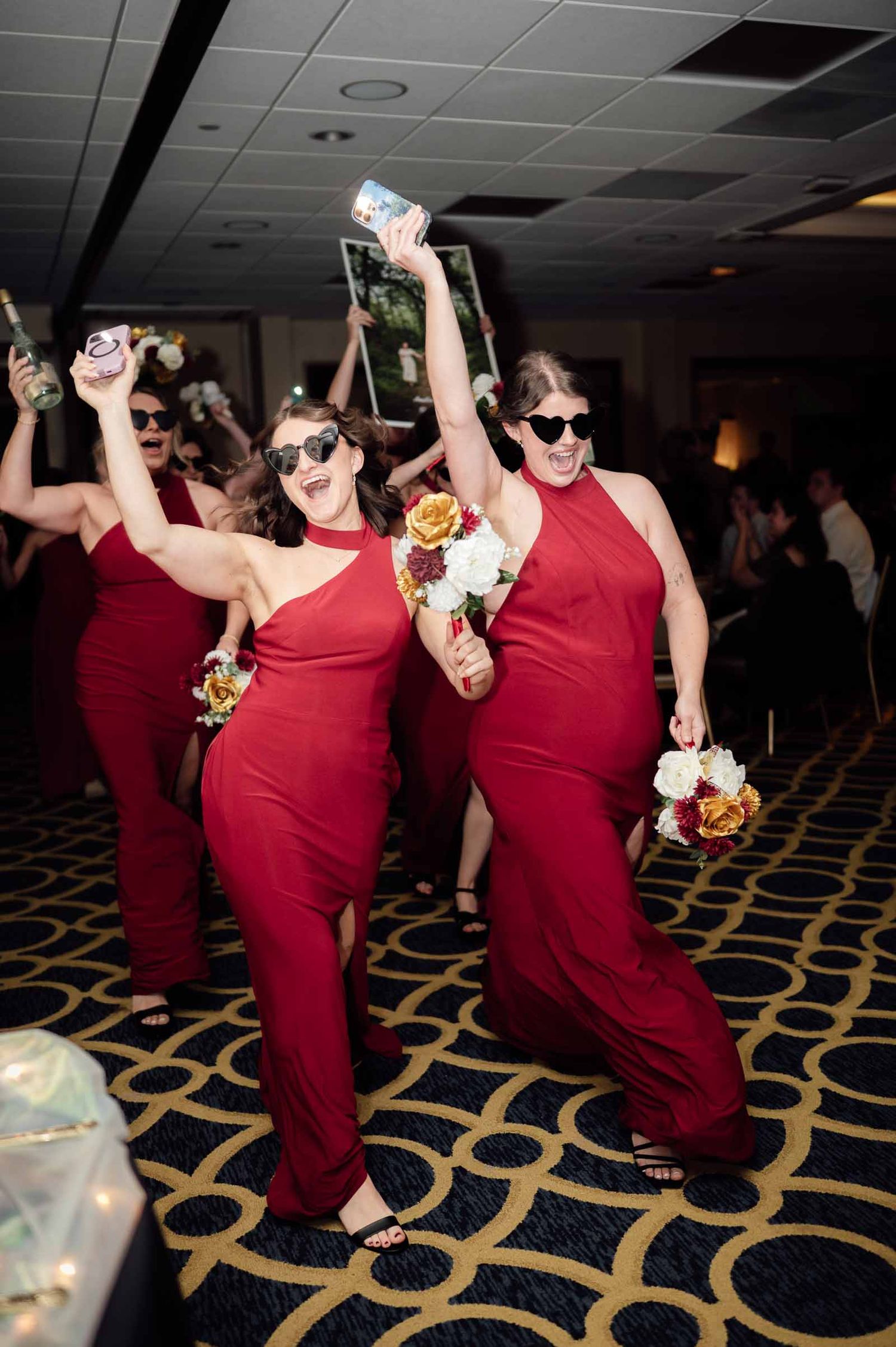 Bridesmaids in red halter gowns dancing at wedding reception, wearing sunglasses and holding bouquets.
