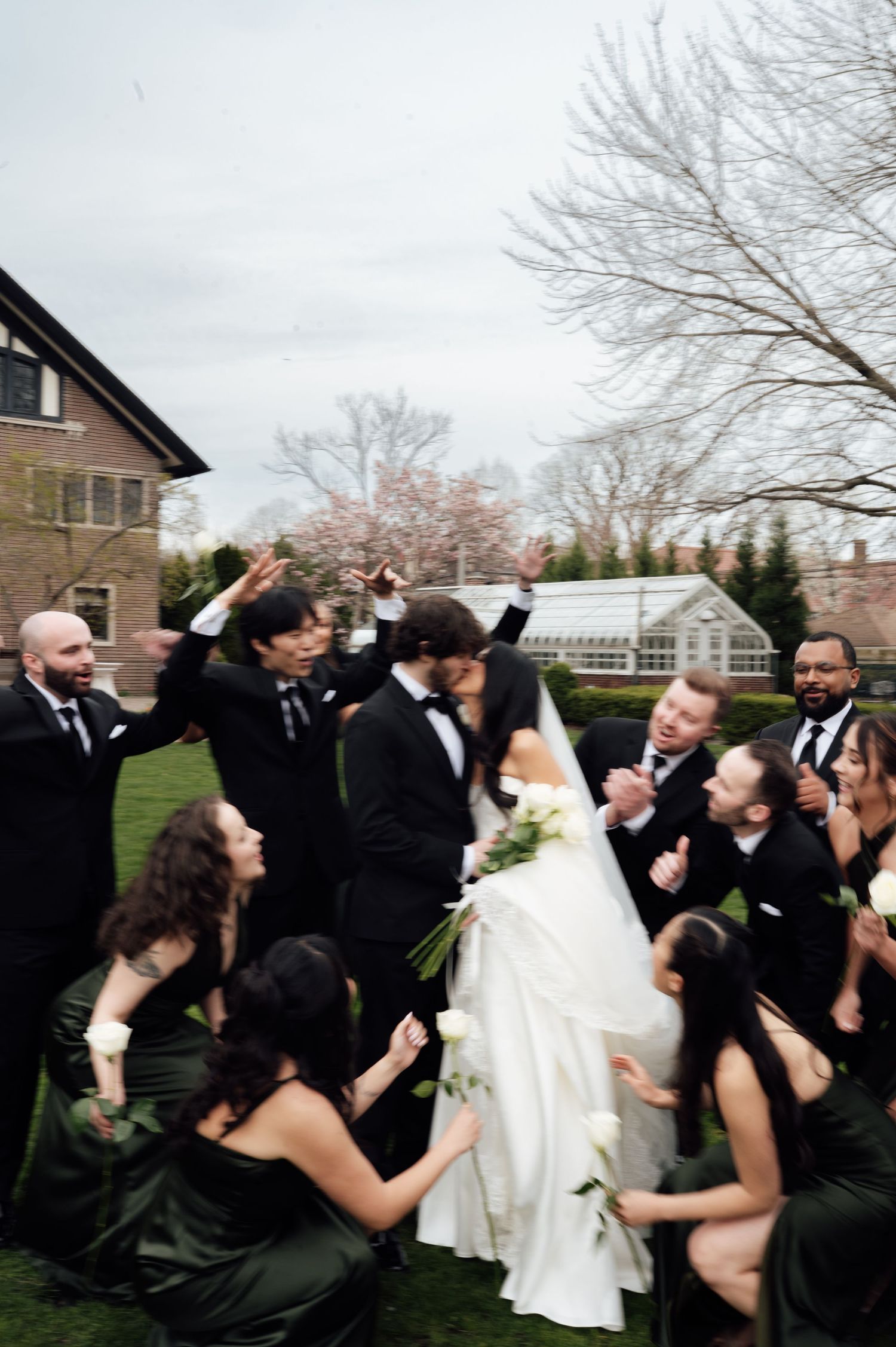 Bride and groom share a kiss surrounded by cheering wedding party in black attire on a green lawn.