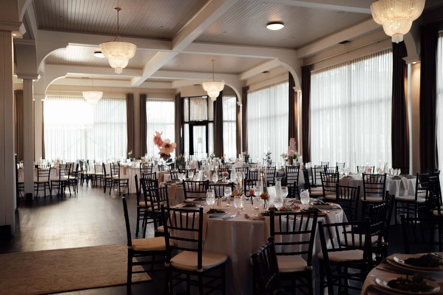 Elegant banquet hall with round tables, chiavari chairs, white linens, chandelier lighting, and large windows for a wedding reception.