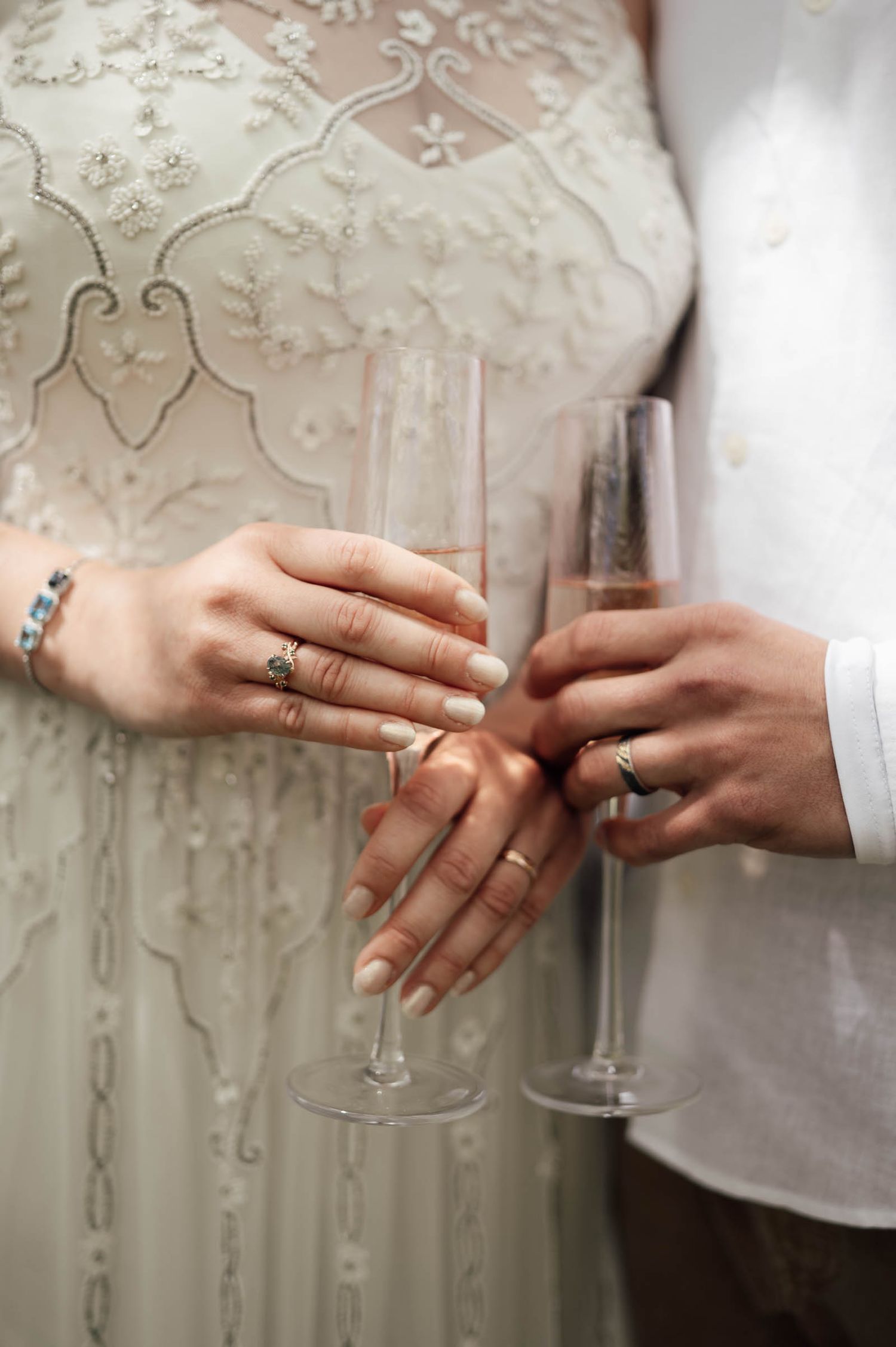 Bride and groom holding champagne flutes, showcasing wedding rings on their hands, wearing elegant white wedding attire.