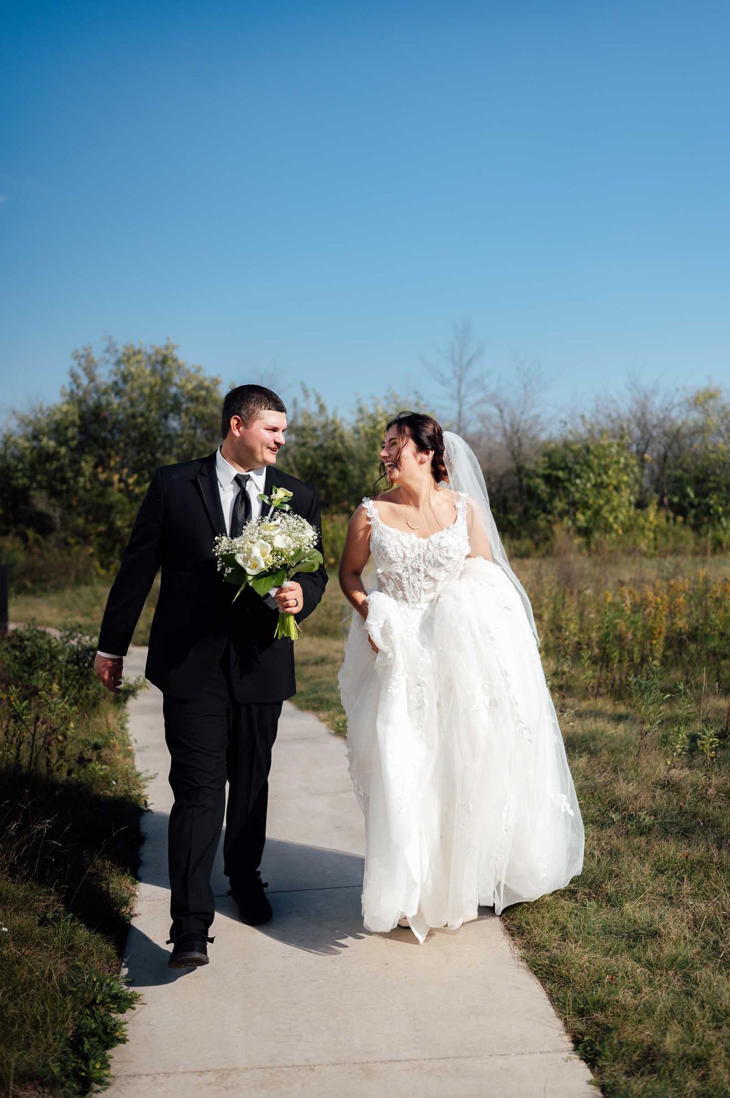Smiling bride and groom walk hand-in-hand on a sunny path, bride in white gown holding bouquet.