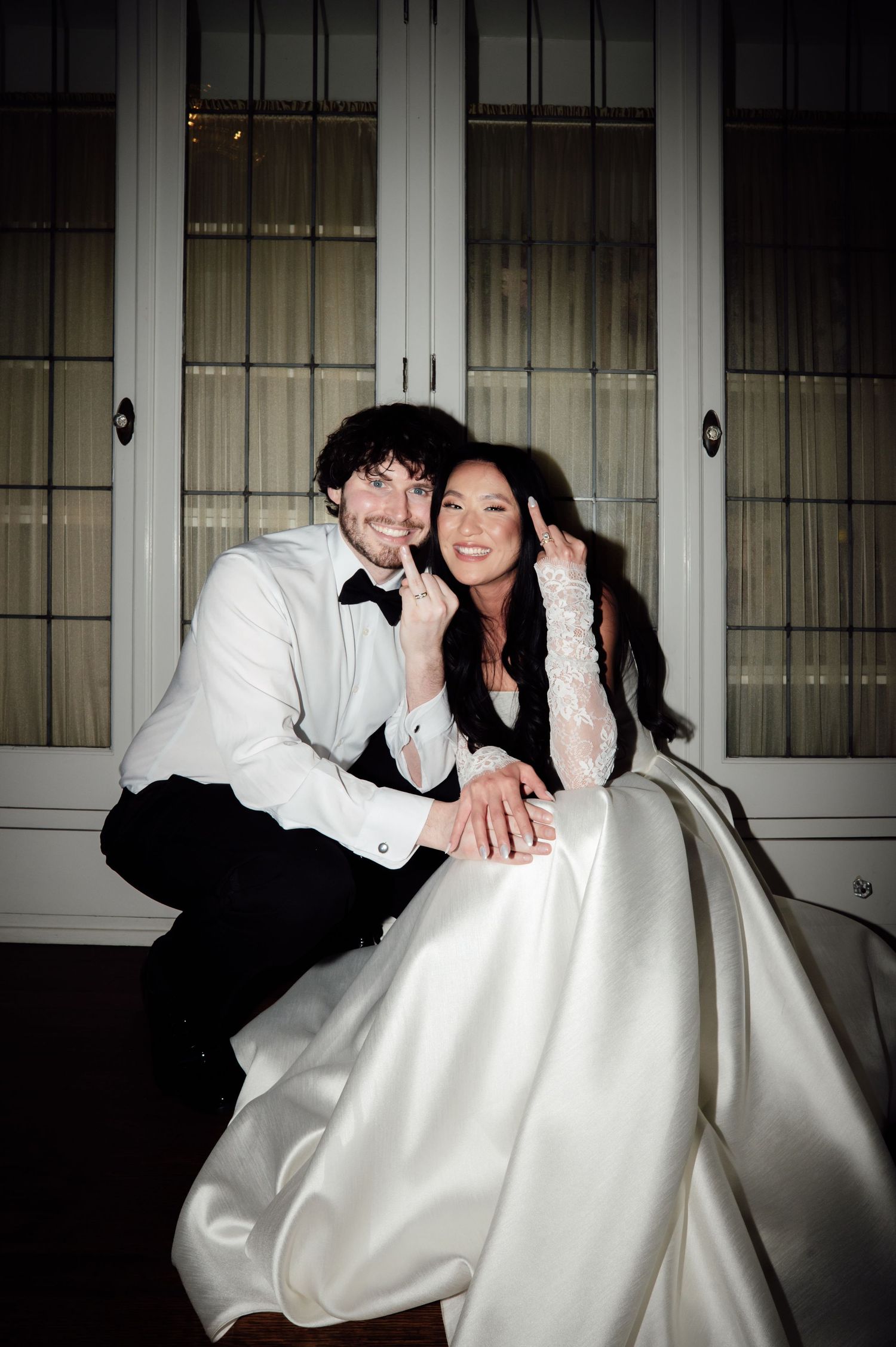 Happy couple in wedding attire poses together, bride in white gown with lace sleeves and groom in black tuxedo.