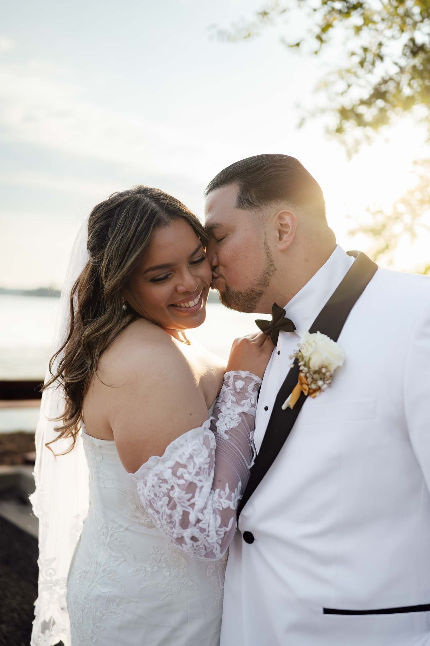 A groom in a white tuxedo kisses his smiling bride in a lace off-shoulder gown during a golden hour outdoor wedding.