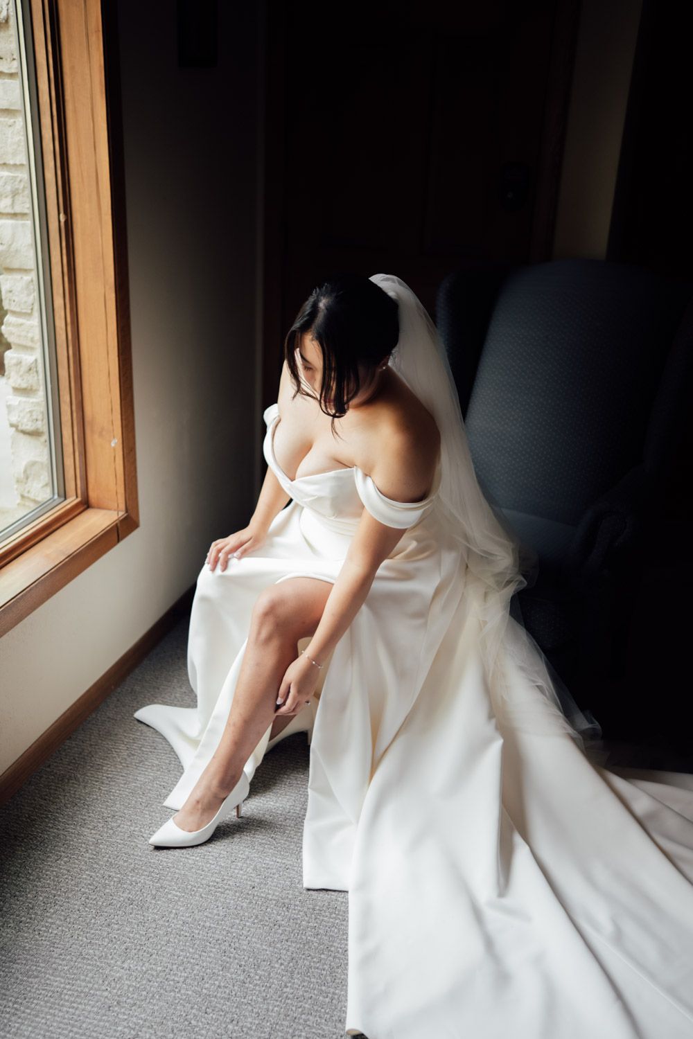 Bride in off-shoulder white wedding gown and veil sits by window, adjusting her white heels before ceremony.