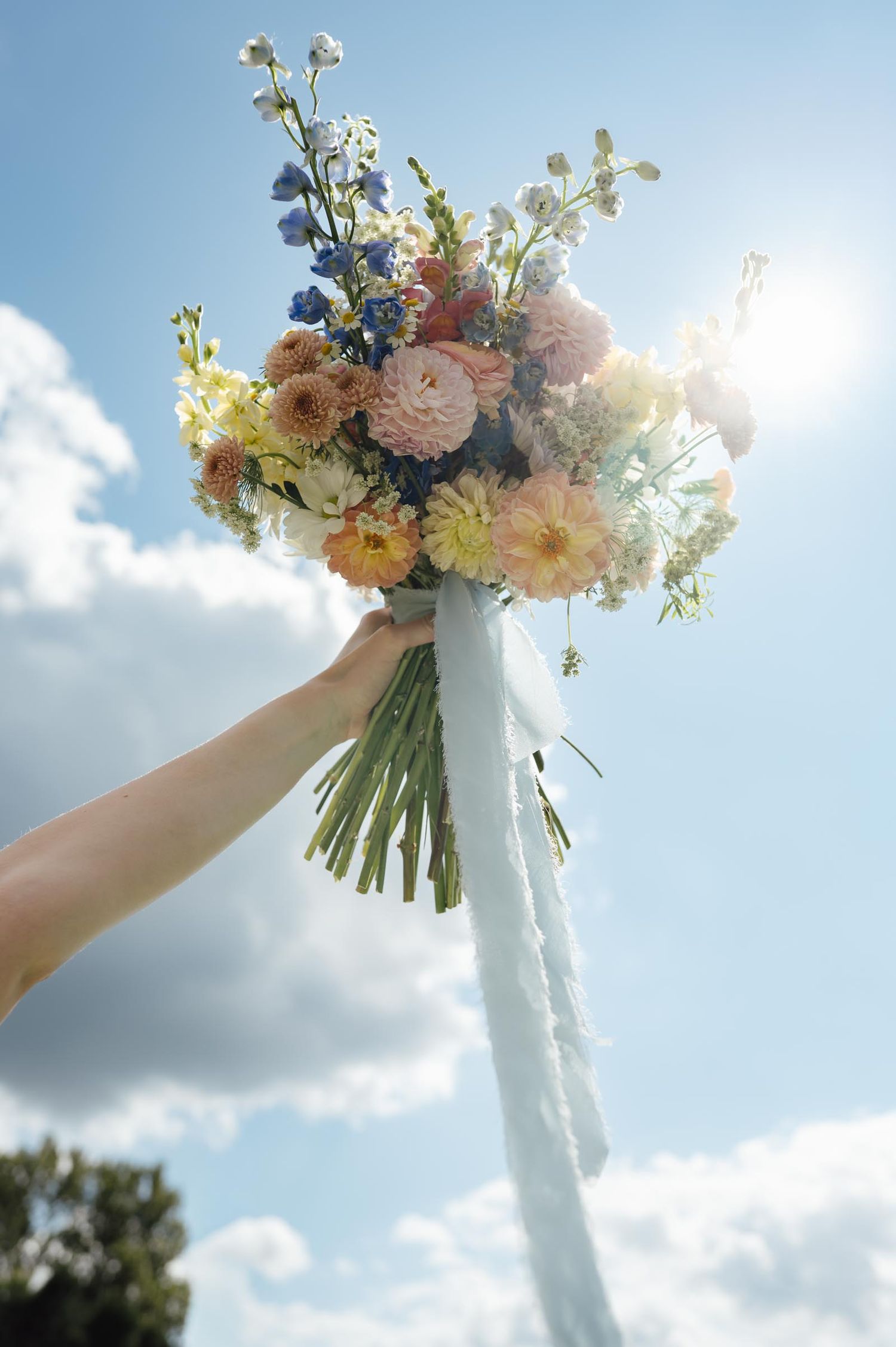 A hand holds a colorful wedding bouquet of dahlias and wildflowers with white ribbons against a sunny blue sky.