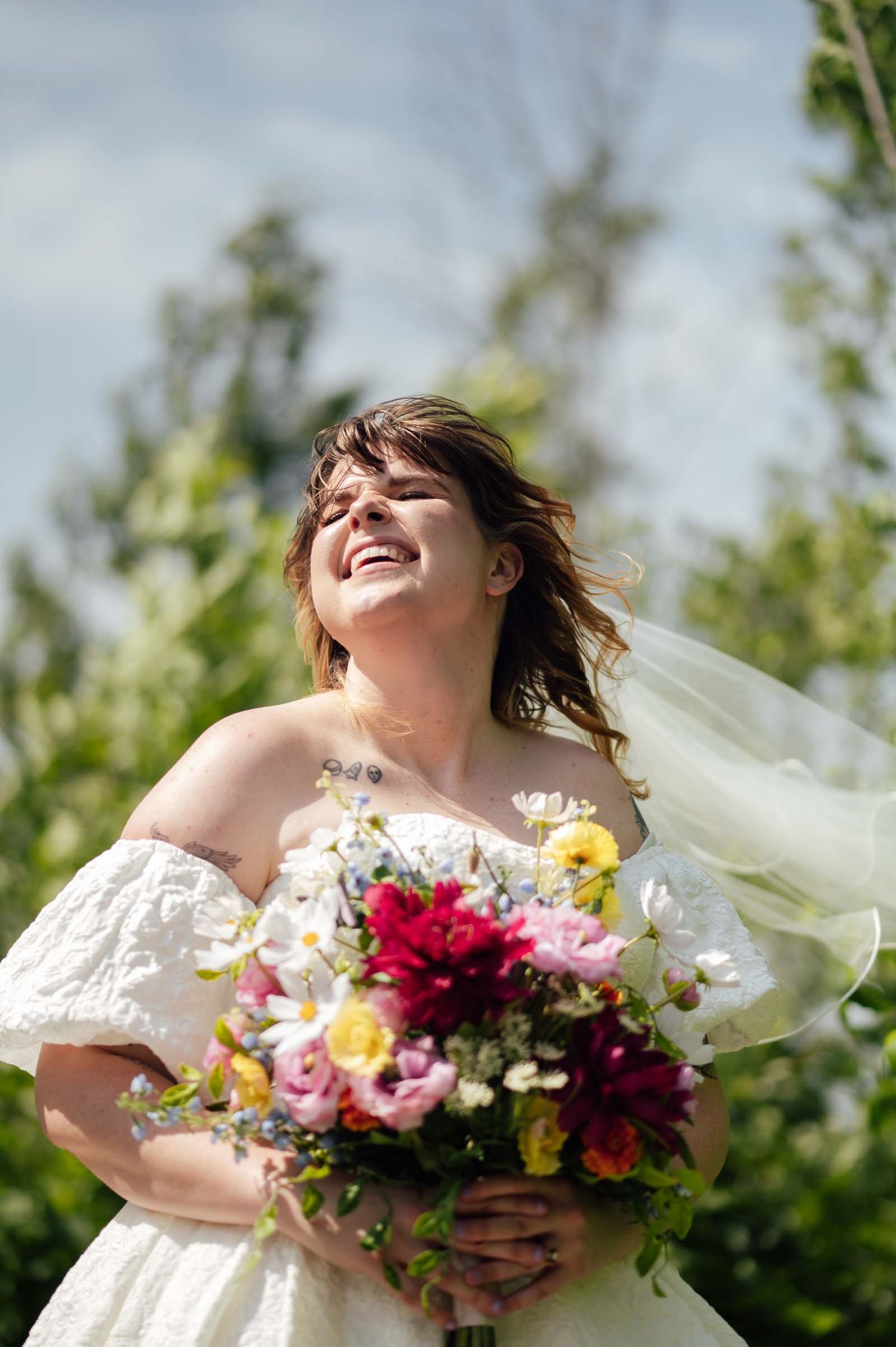 Joyful bride in off-shoulder white gown holds colorful bouquet outdoors on a bright sunny day.