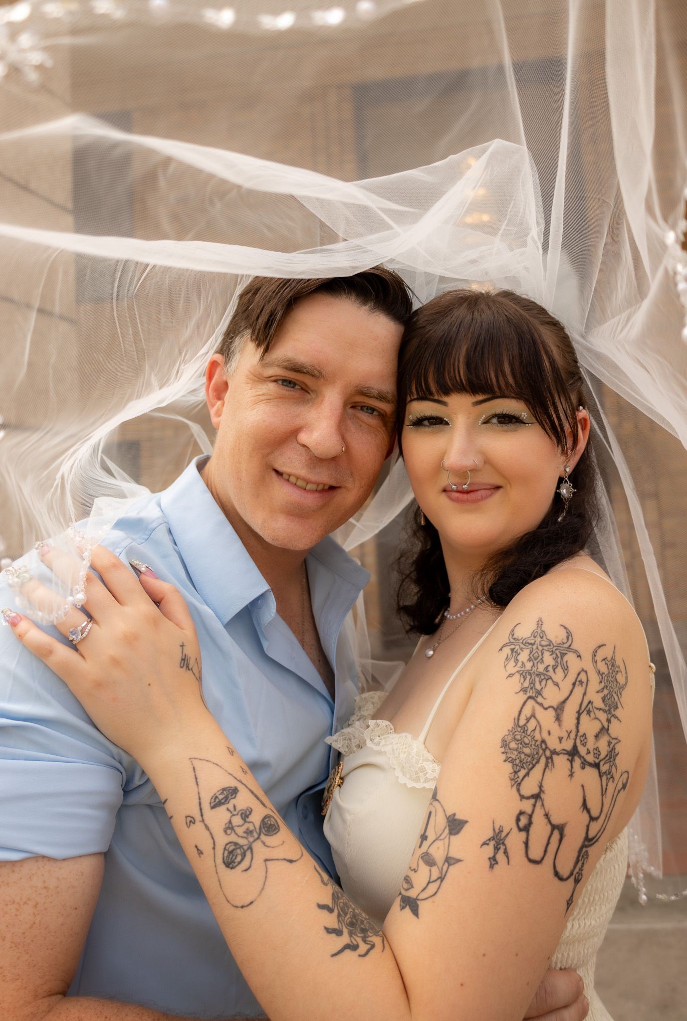 A couple poses together under a sheer veil, showing off matching tattoo designs on their arms.