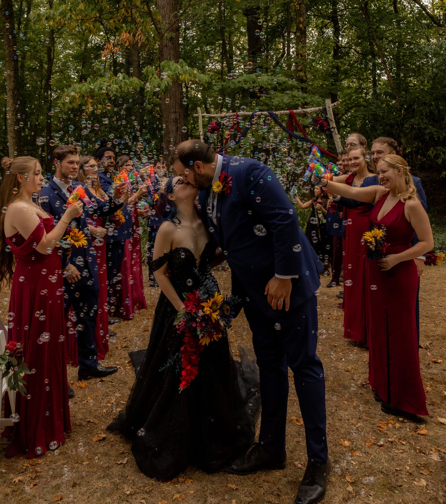 A group in burgundy dresses and dark suits gathers outdoors among autumn trees for a wedding celebration.