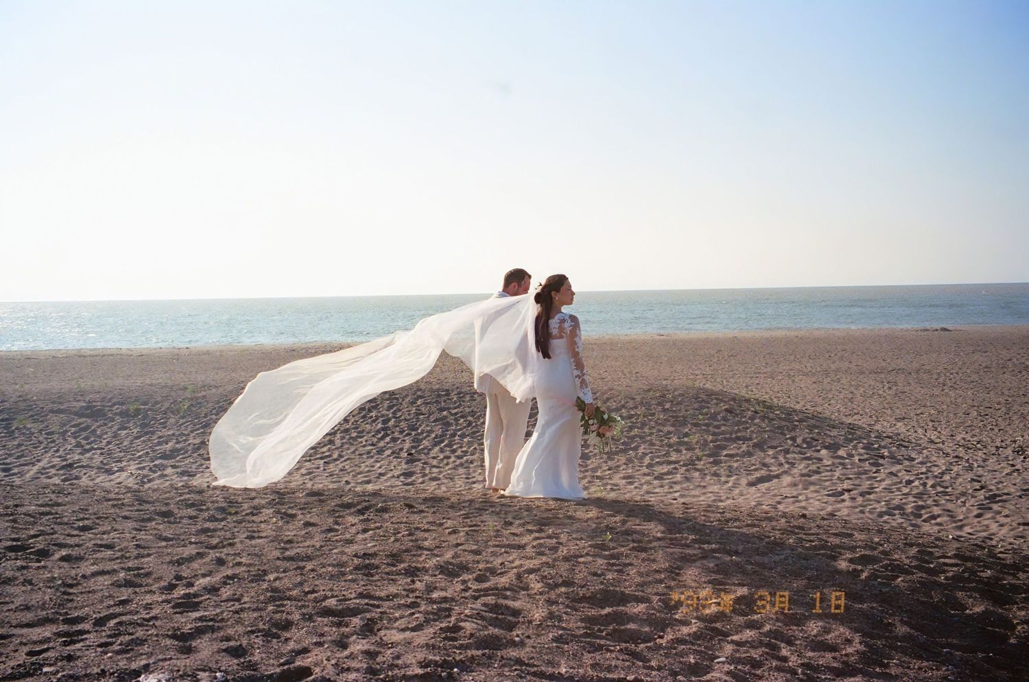 A white wedding dress flows in the wind on a scenic beach as a couple embraces during their seaside photo session.