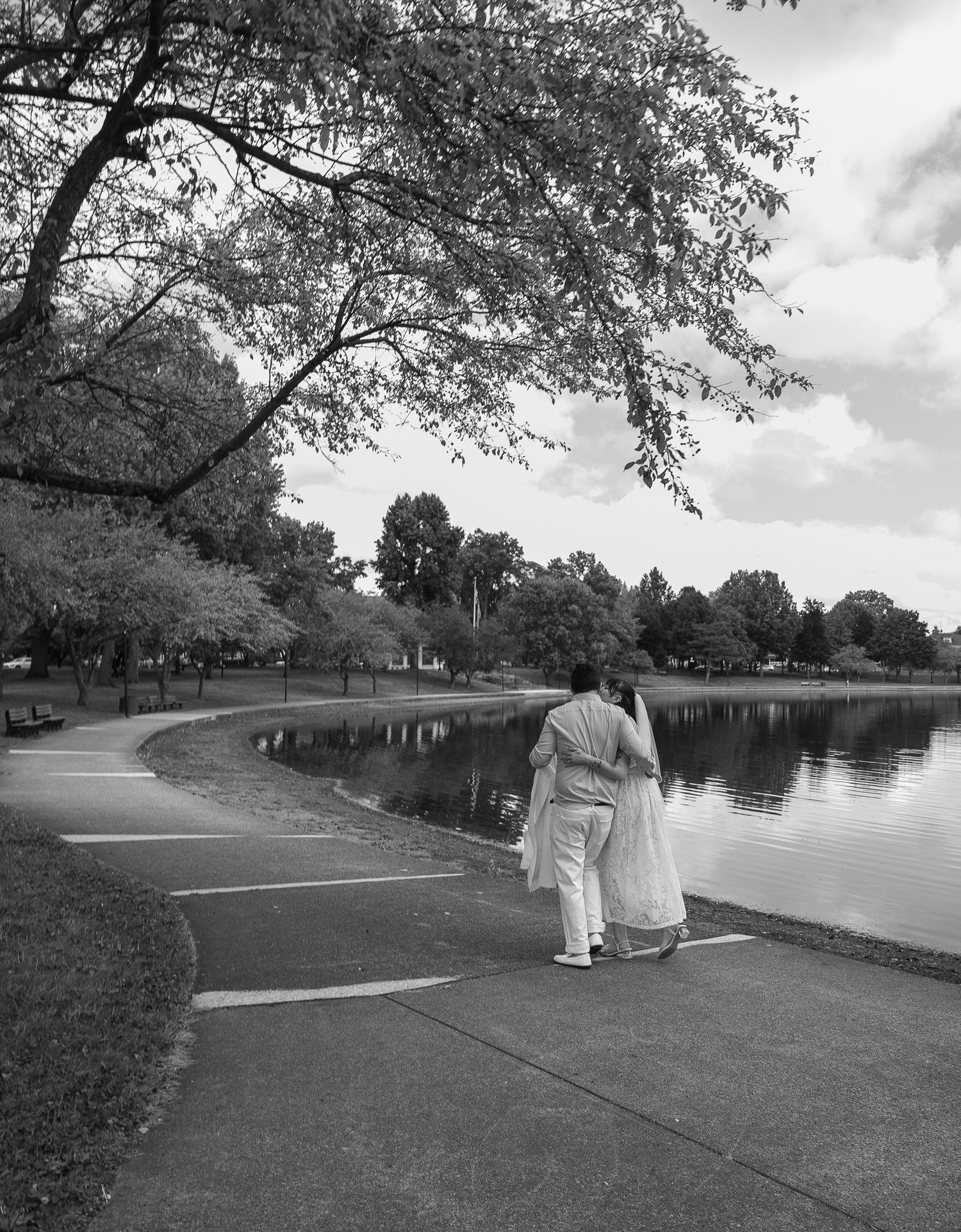 Two people walking along a peaceful lakeside path under overhanging trees on a cloudy day.