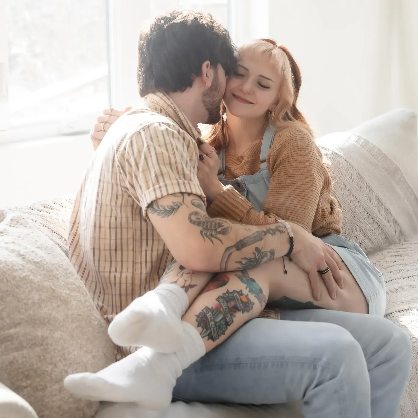 Intimate couple sharing a tender moment on a couch, with visible tattoos and casual clothing.